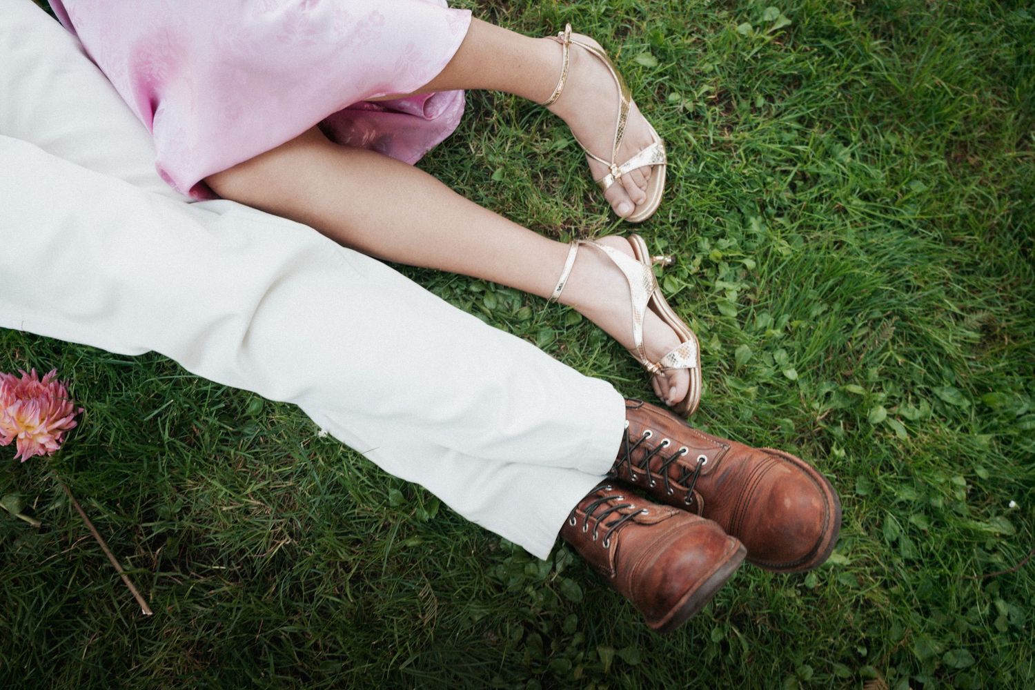 Bride and groom sitting on green grass, wearing sandals and brown leather boots beside a pink flower.