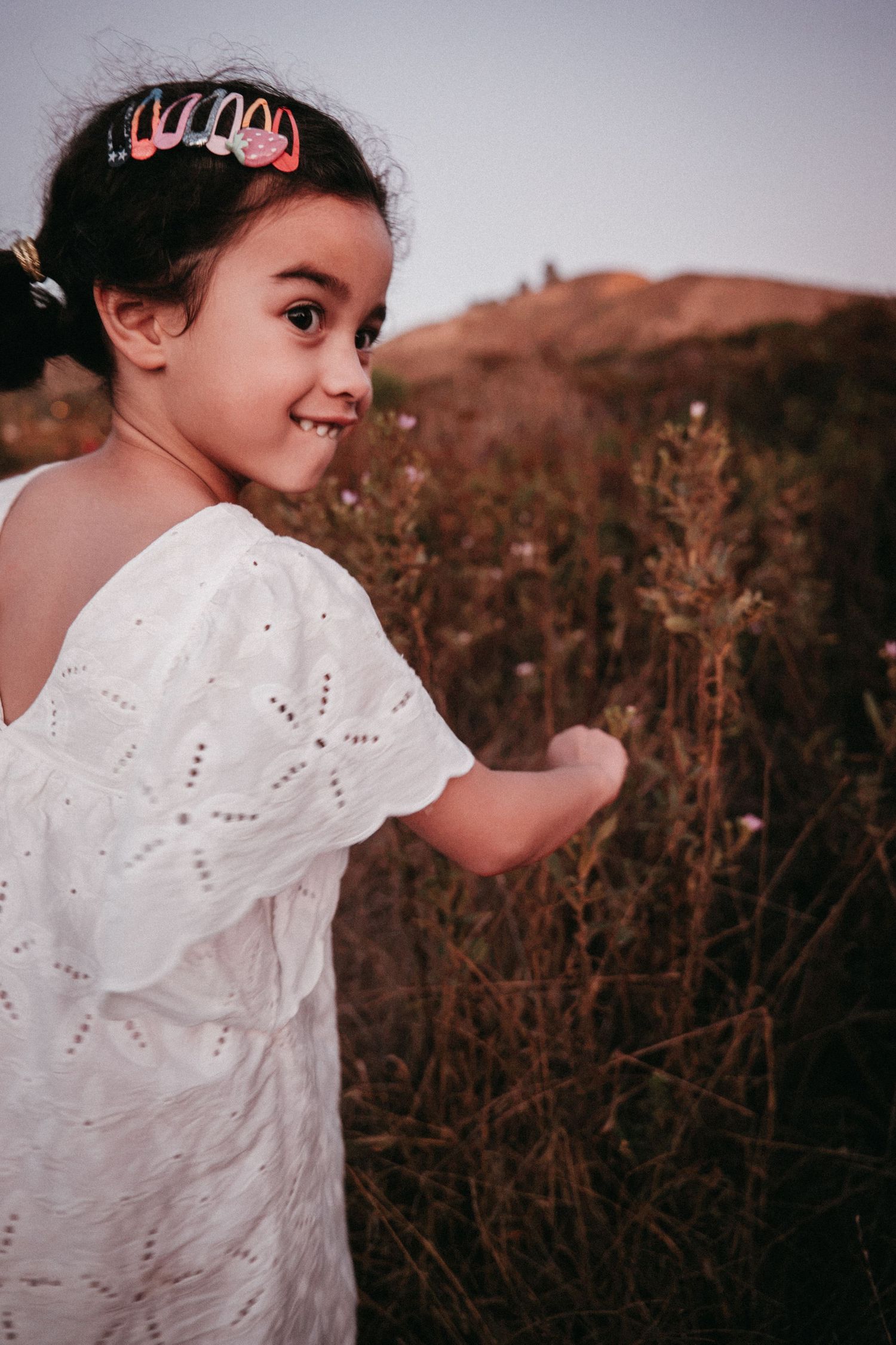 A young girl in a white dress with hair clips smiles outdoors in a field at dusk.