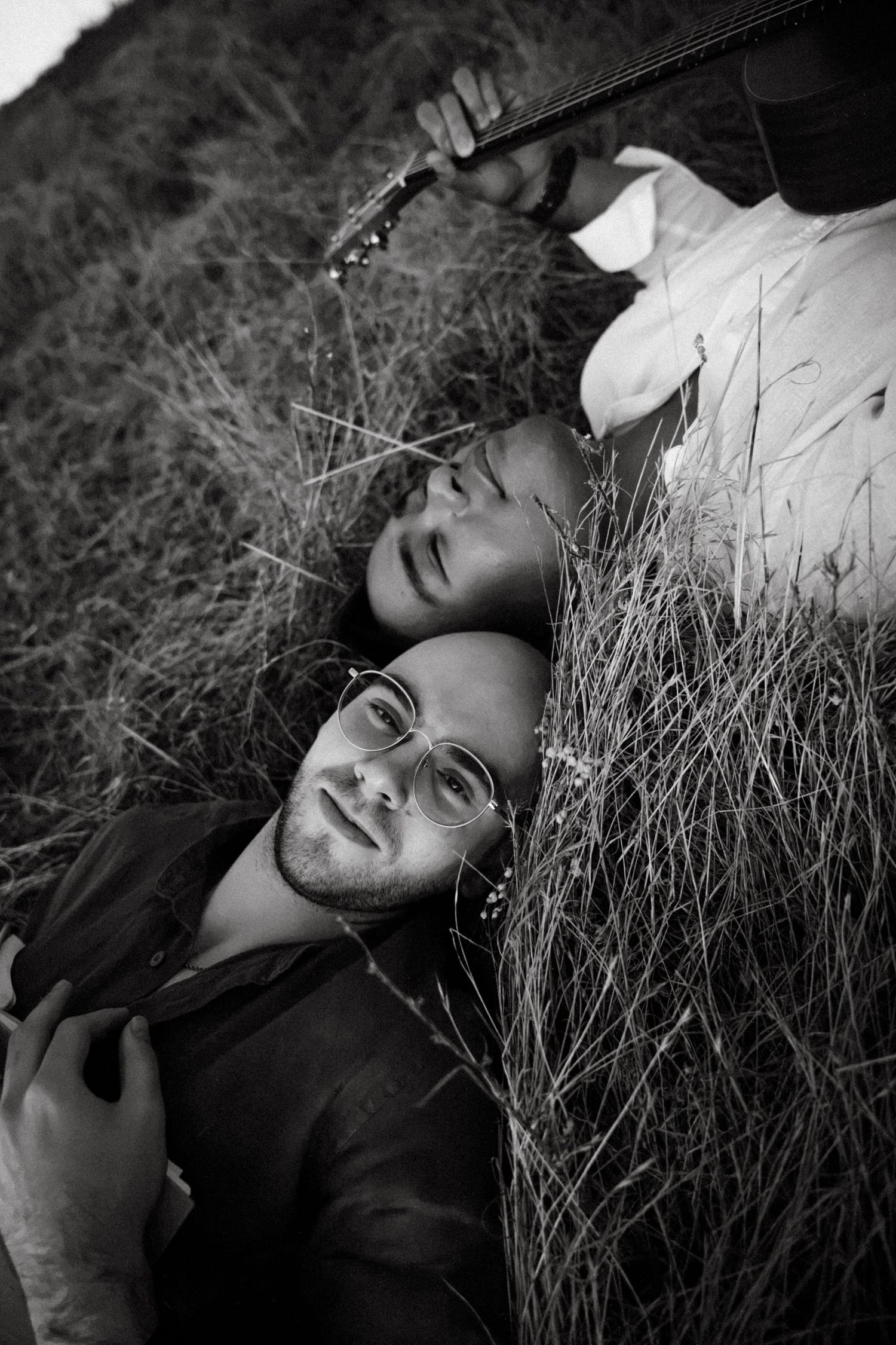 Two men lying in tall grass, one wearing glasses, in a romantic black and white outdoor portrait.