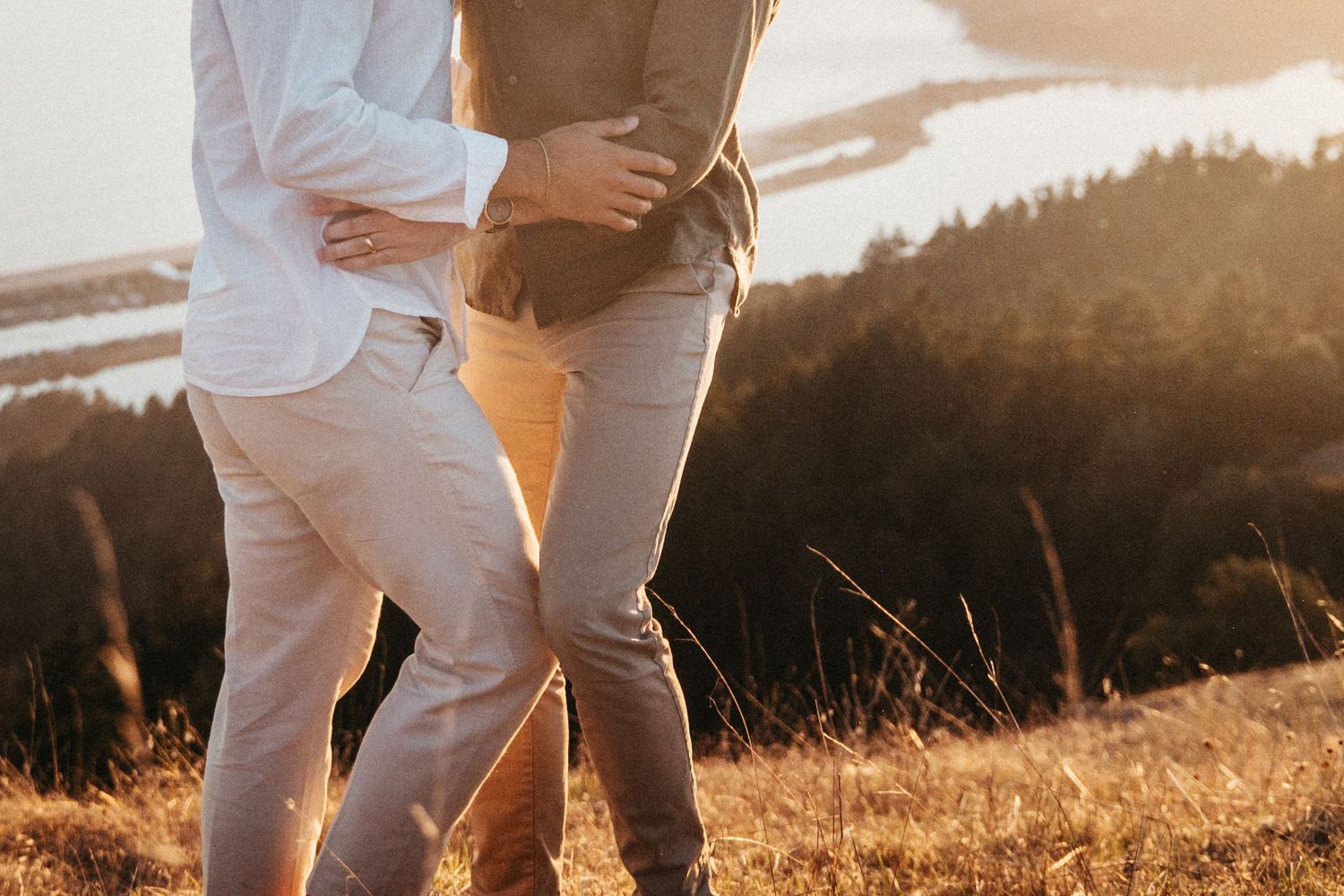 Couple embracing on a golden hillside at sunset with a scenic river valley in the background.