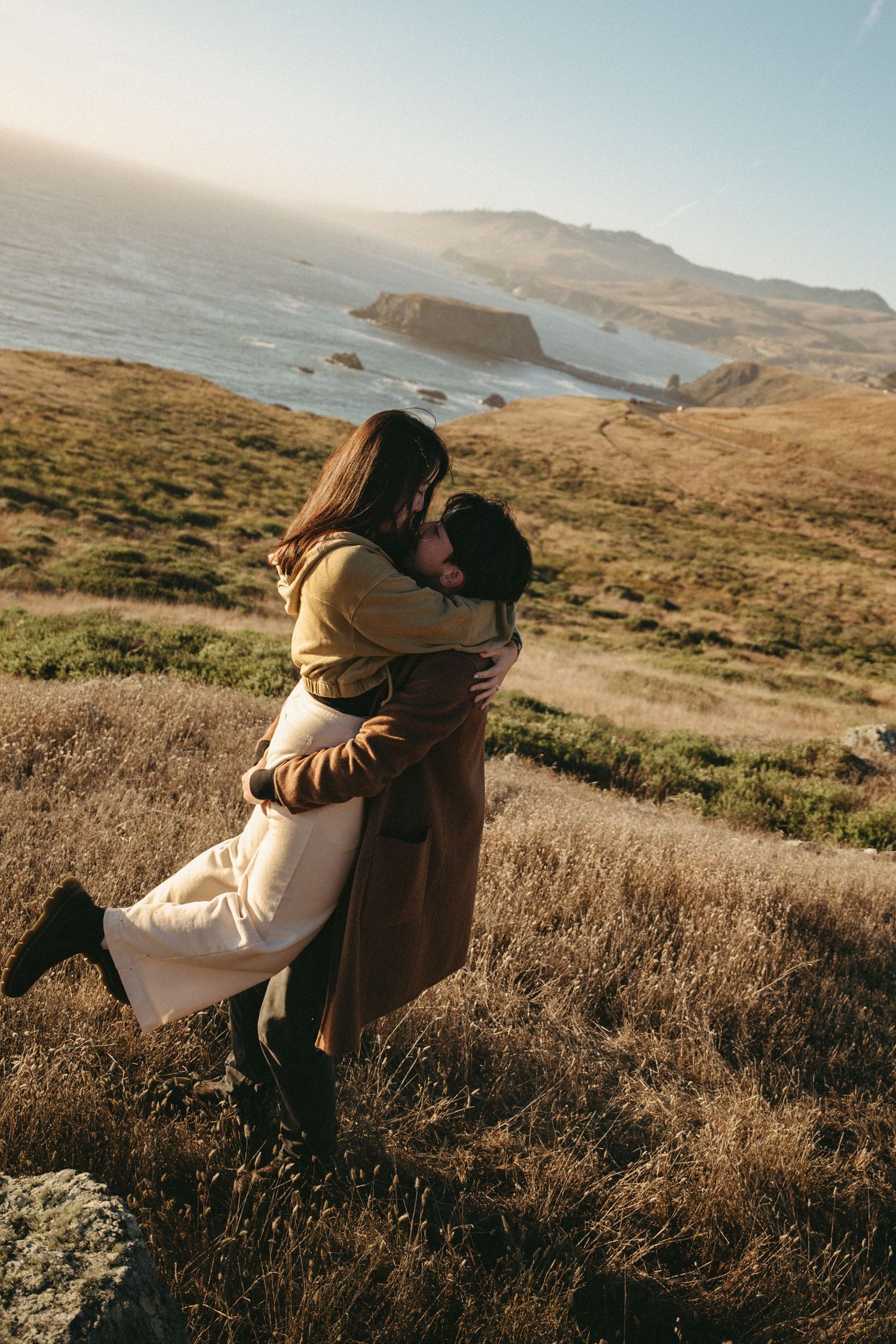 Couple embracing in a romantic lift on a grassy coastal hillside with ocean waves and rugged cliffs in the golden-hour background.