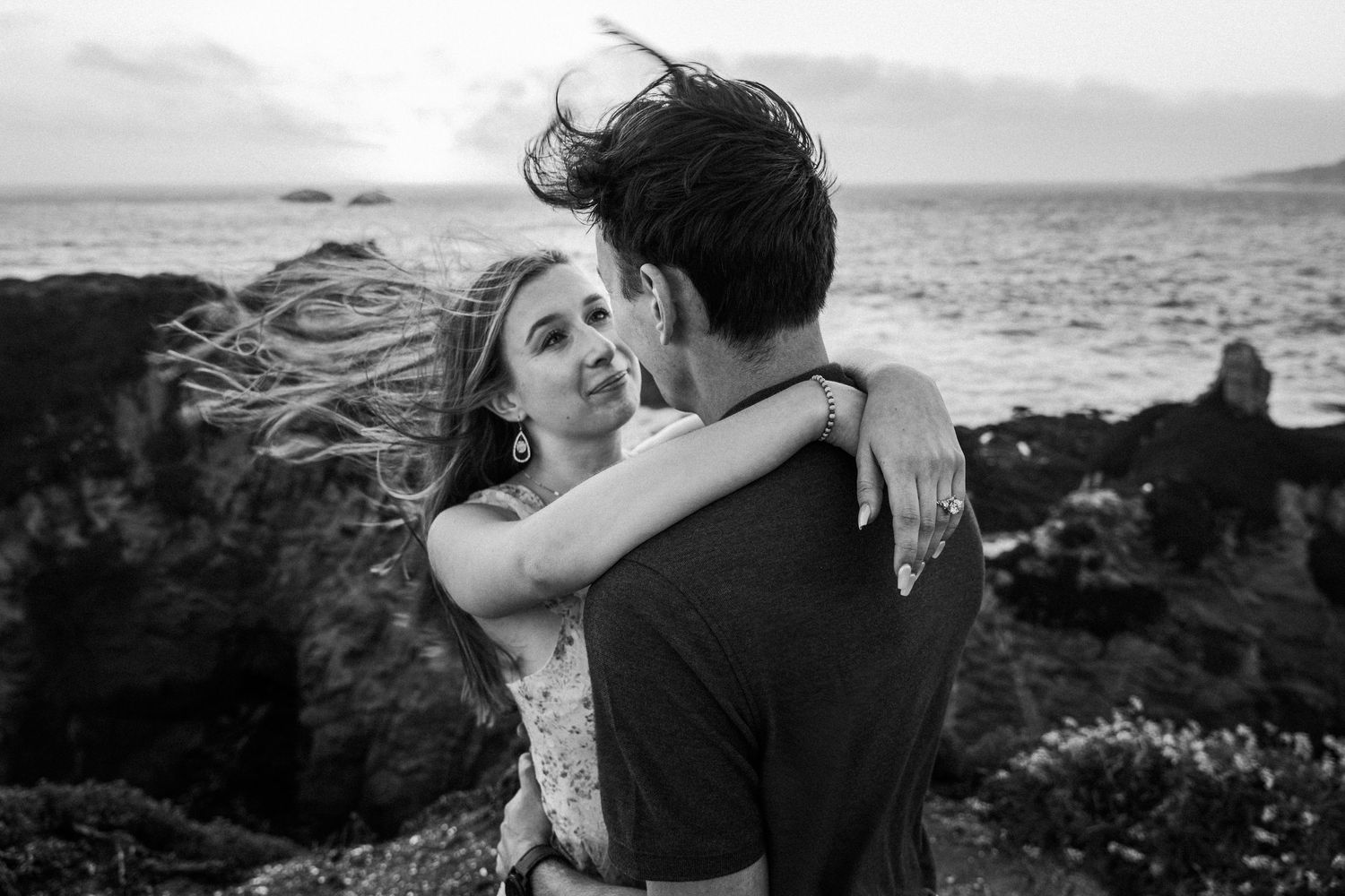 Black and white photo of a couple embracing near rocky ocean cliffs at sunset, hair blowing in wind.