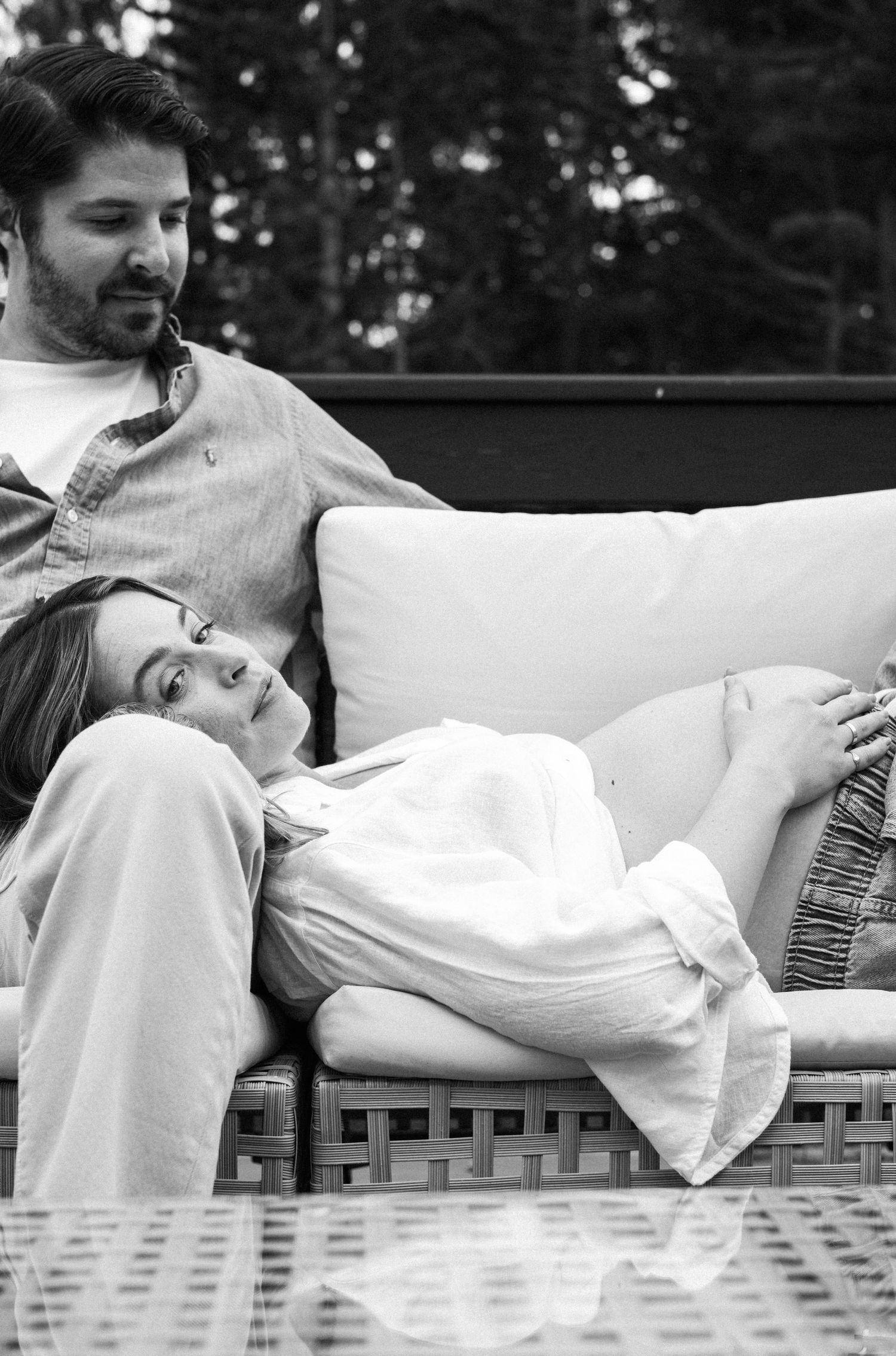 Black and white photo of a pregnant woman resting on outdoor sofa with her partner during maternity shoot.
