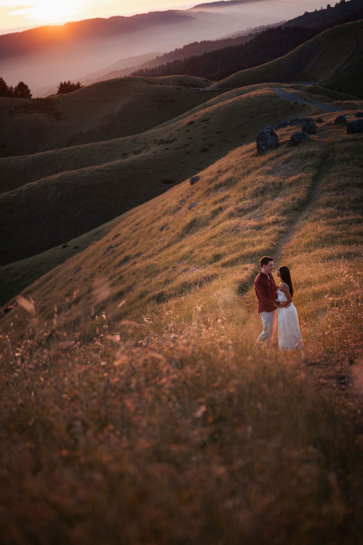 Romantic couple embracing at sunset on rolling golden hills with foggy mountains beyond.