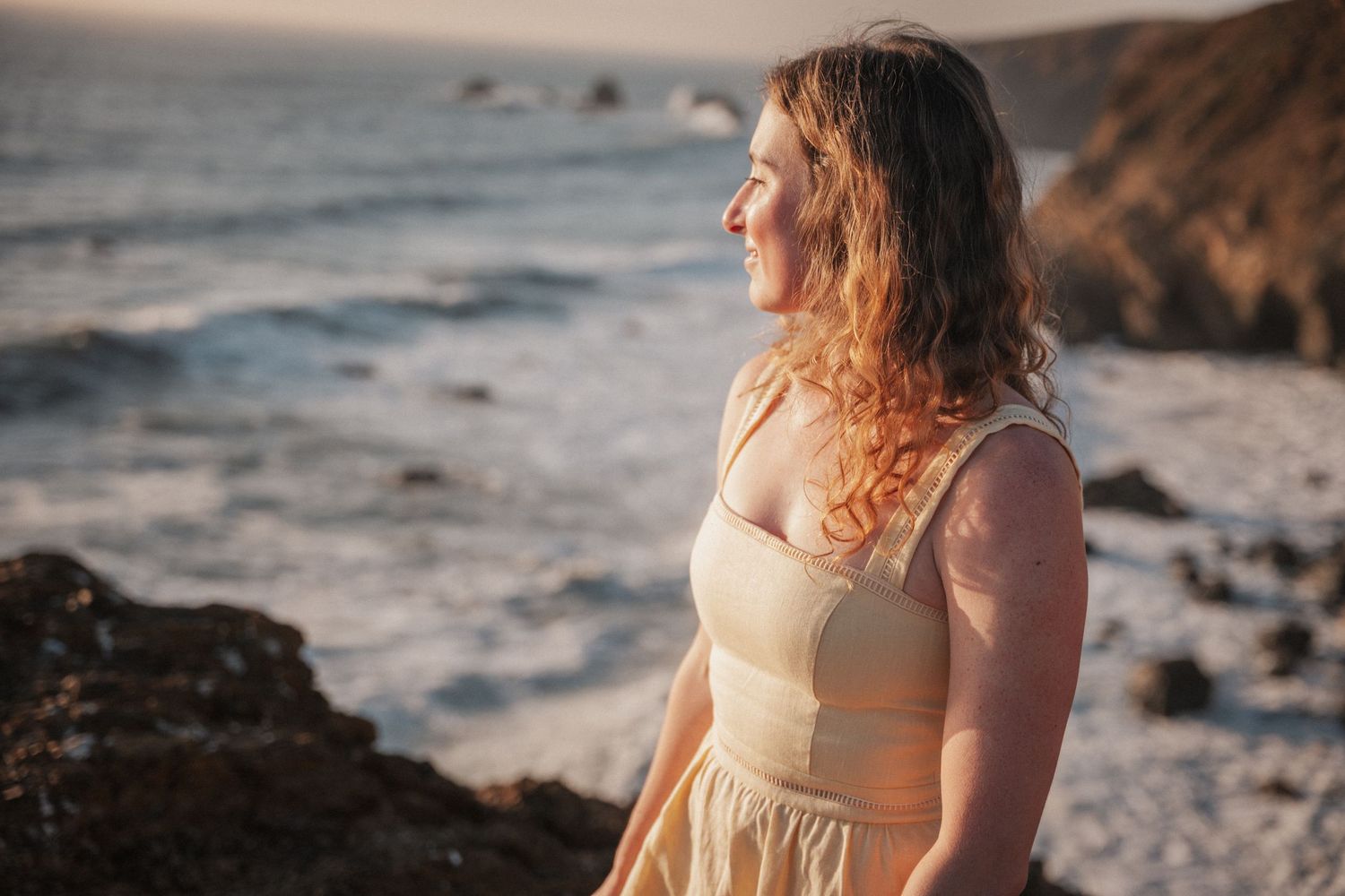 A woman with curly red hair in a cream dress gazes at the ocean from rocky coastal cliffs at golden hour.