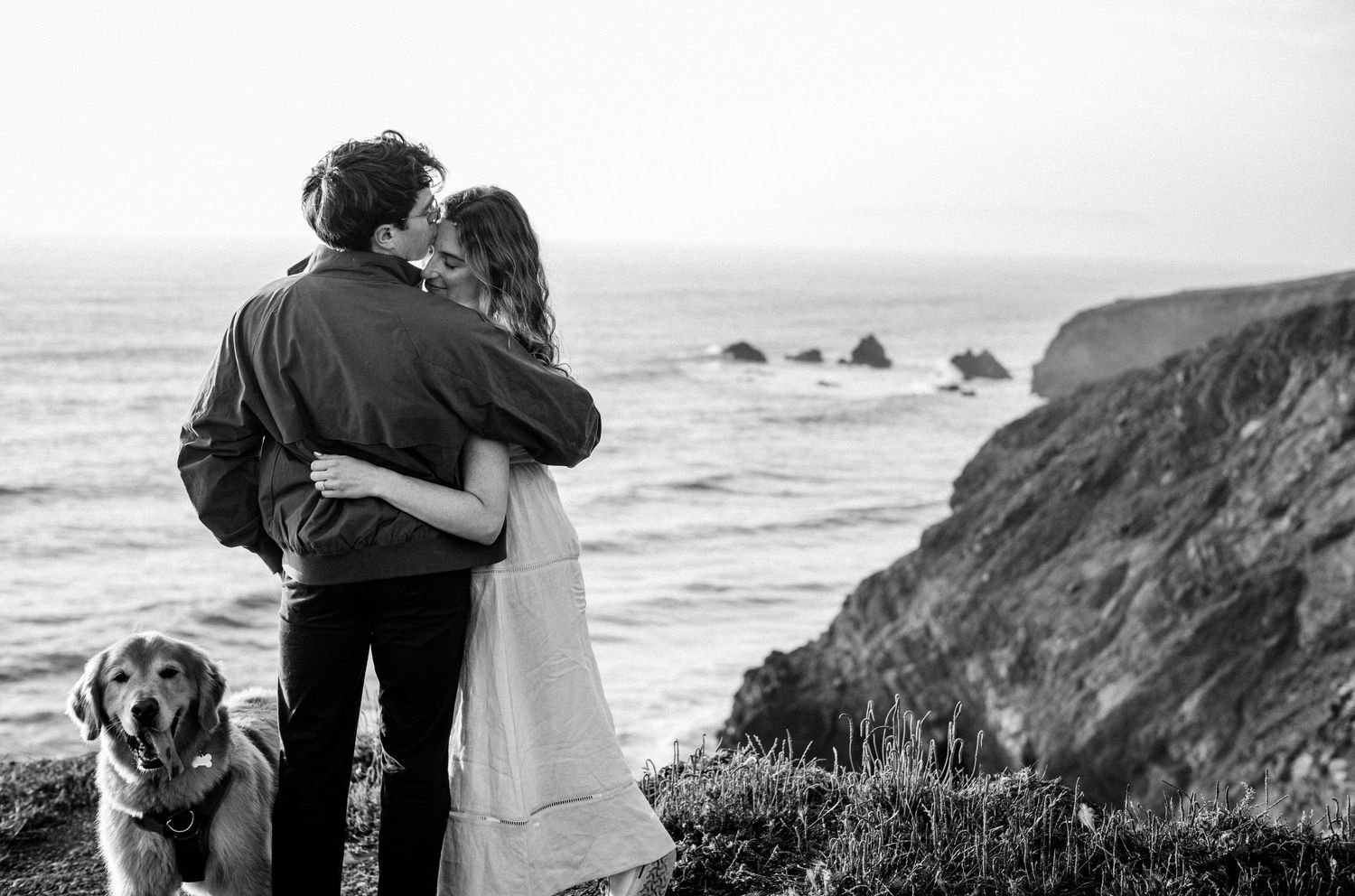 Couple embracing and kissing on a coastal cliff with a golden retriever dog, ocean and rocks in black and white.
