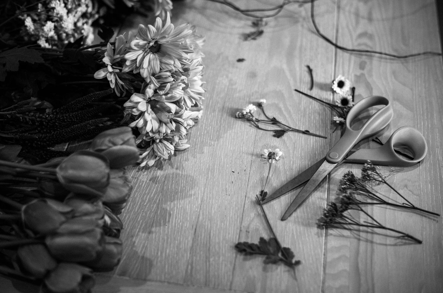 Black and white flat lay of wedding flowers, scissors, and ribbon on a wooden floor.