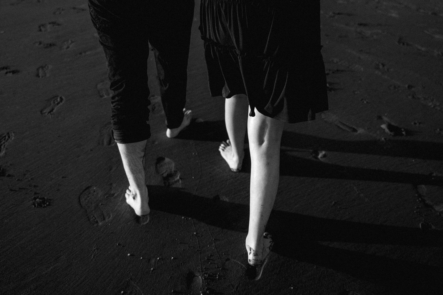 Two barefoot people walking on a dark sandy beach, leaving footprints, captured in black and white.