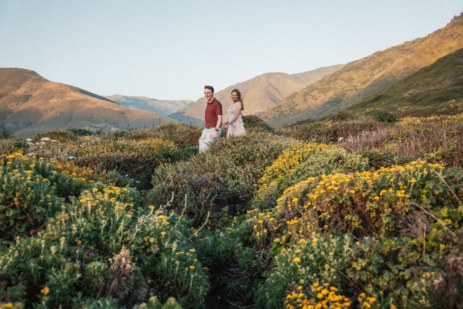Couple standing among yellow wildflowers with scenic mountain landscape in golden hour light.
