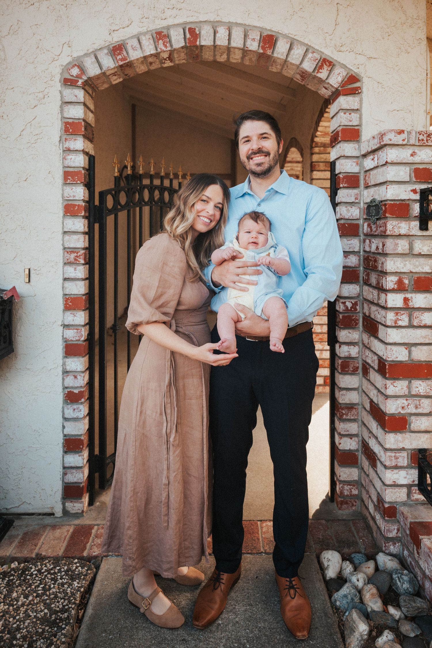 Happy young family posing with their baby in front of a charming brick arched doorway.