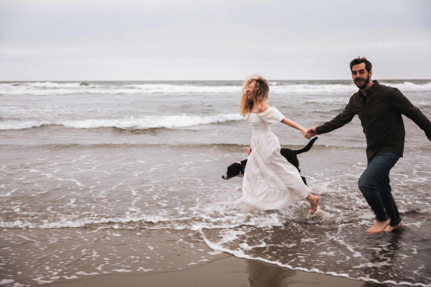 Couple in white dress and dark shirt running barefoot along ocean shoreline on cloudy day.