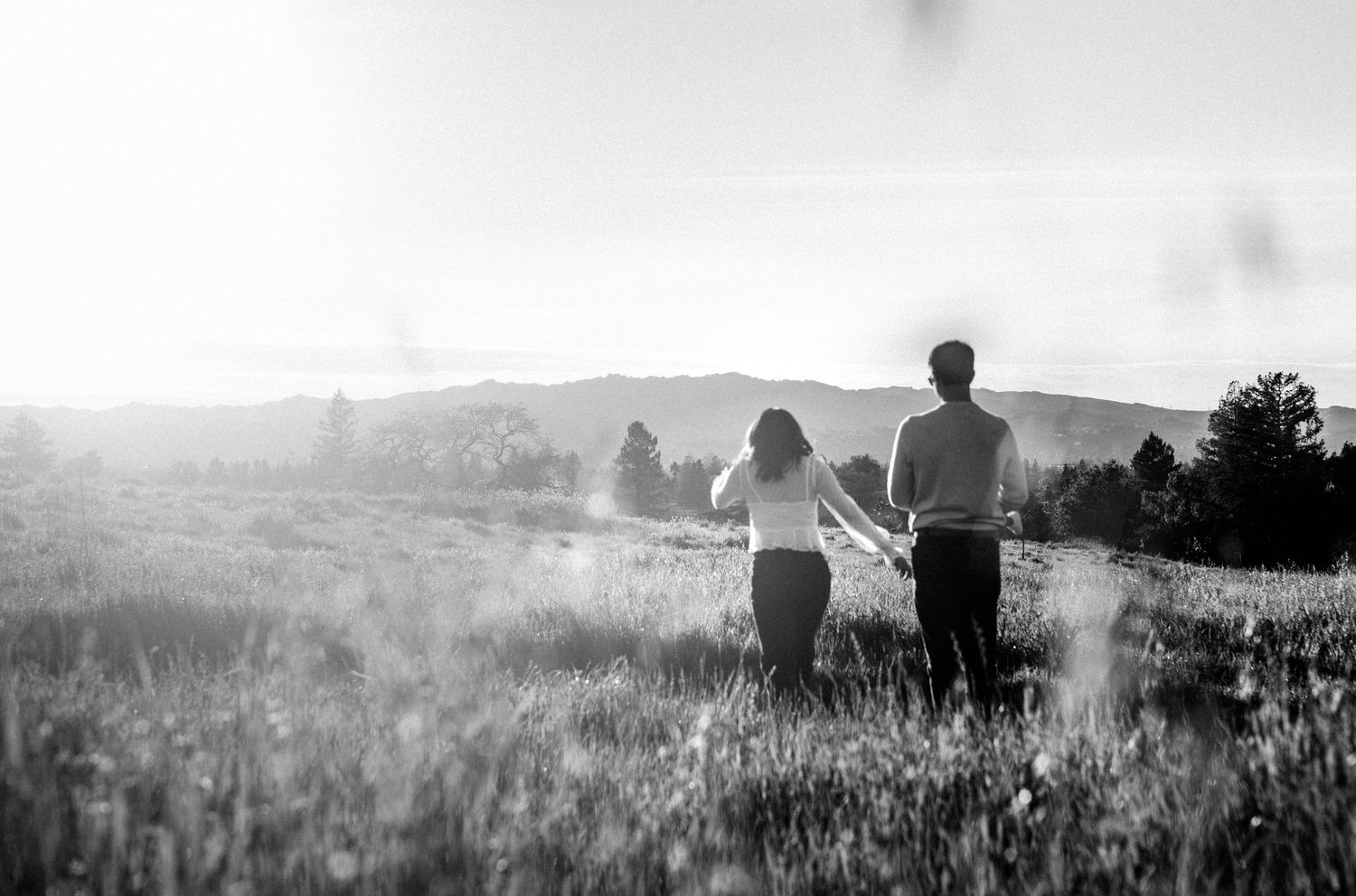 Black and white photo of a couple holding hands walking through a sunlit meadow with mountains in the background.