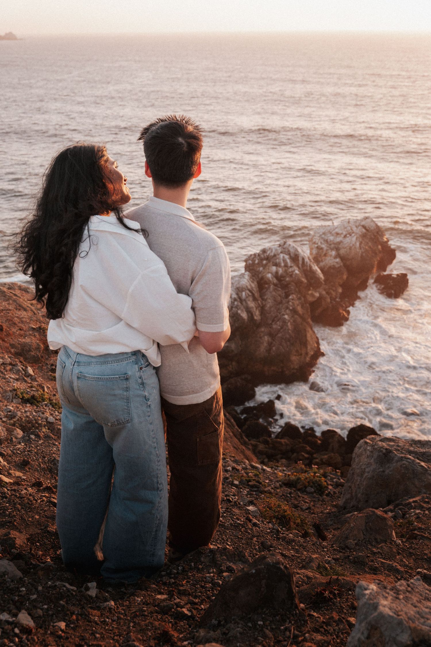 Romantic couple embracing while watching a stunning ocean sunset from a rocky coastal cliff.