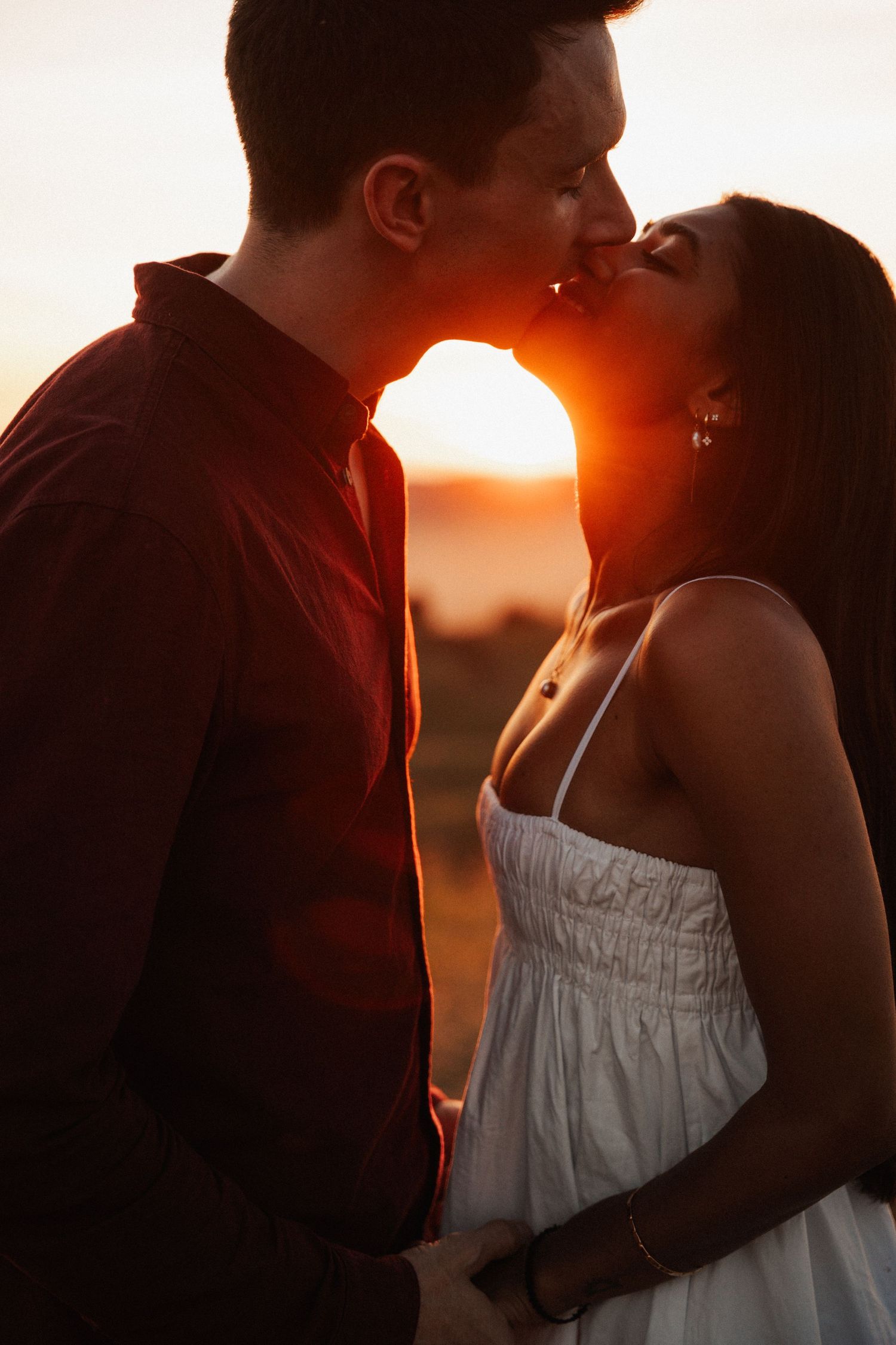 Couple sharing a romantic kiss at golden sunset, woman in white dress, man in red shirt, warm backlit glow.