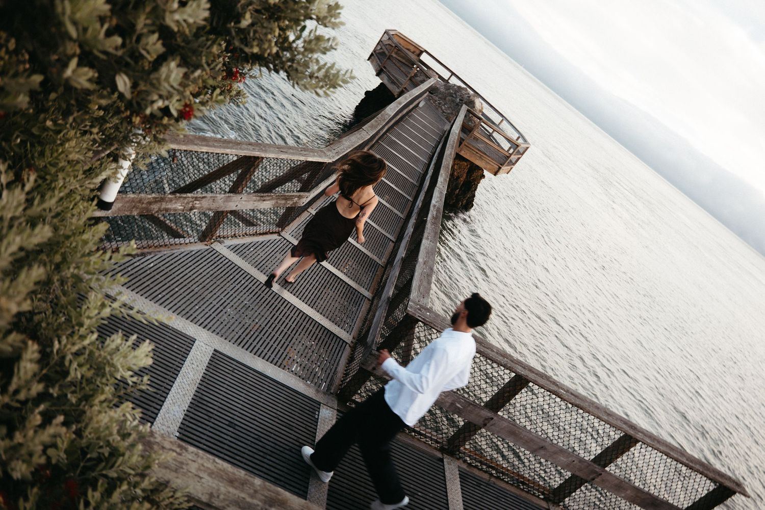 Aerial view of a couple walking up a metal staircase bridge overlooking a calm lake surrounded by trees.