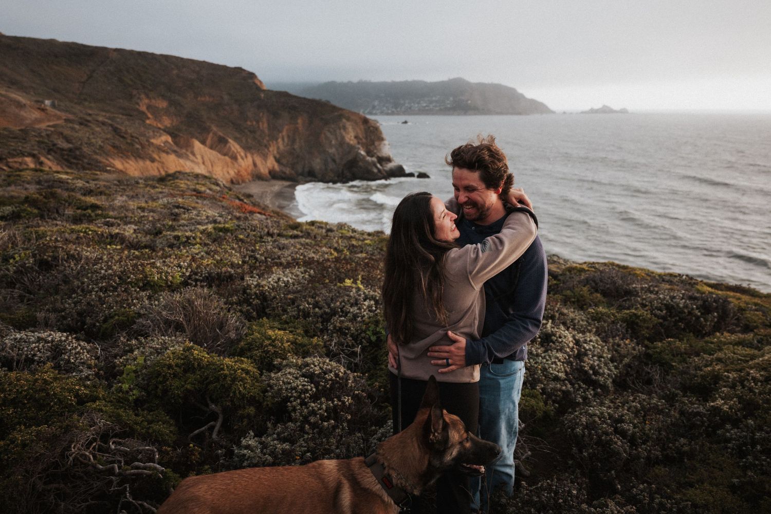 Couple embracing on a coastal cliff with ocean views and a dog nearby on a cloudy day.