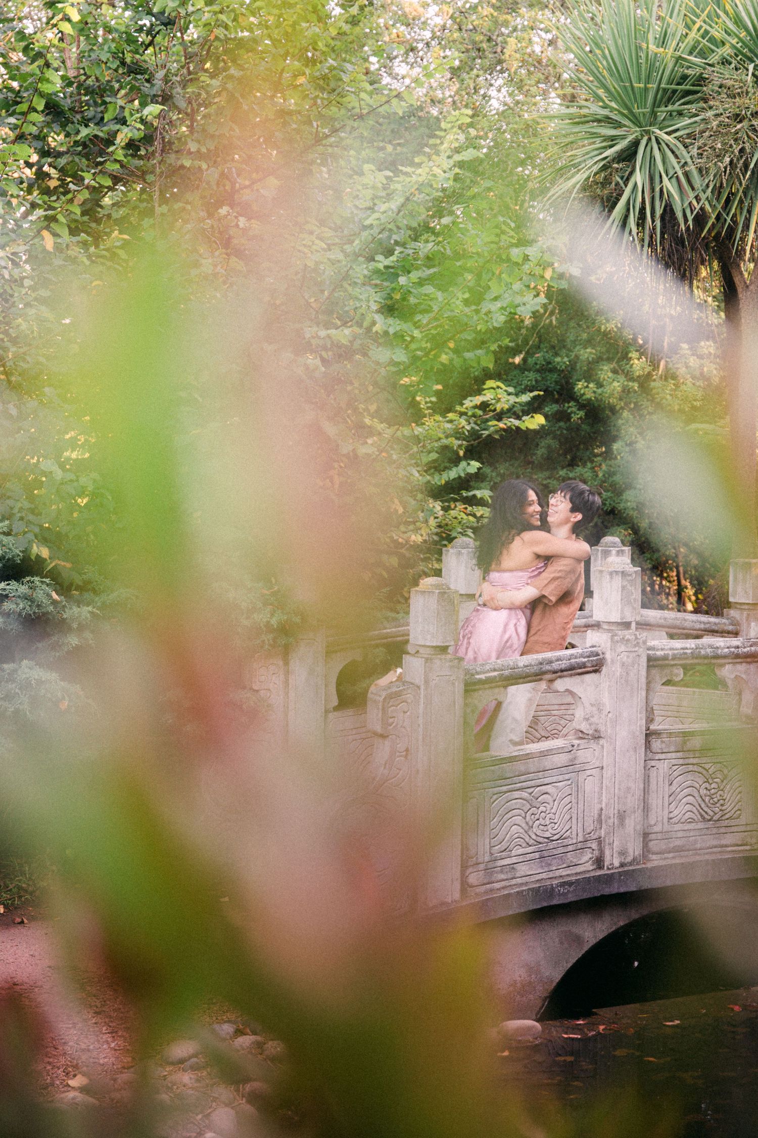 Couple embracing romantically on a stone bridge surrounded by lush green garden foliage.