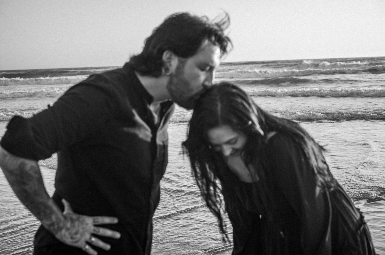 Black and white photo of a couple standing in ocean waves on a beach, man with tattoos leaning toward woman.
