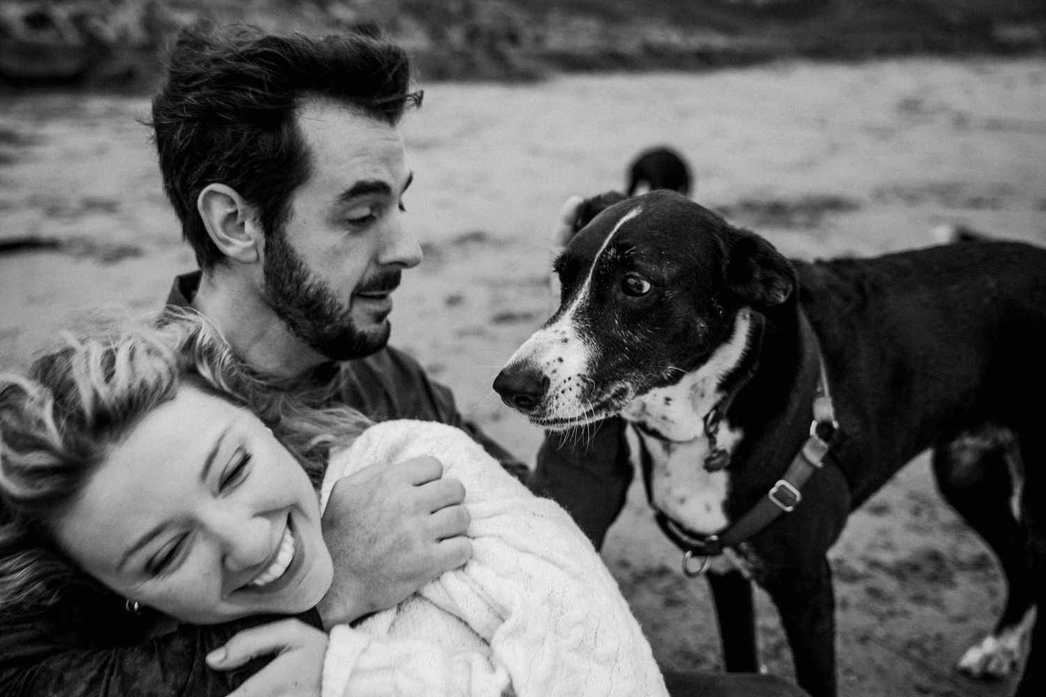 Black and white photo of smiling couple cuddling with their black and white dog on beach.