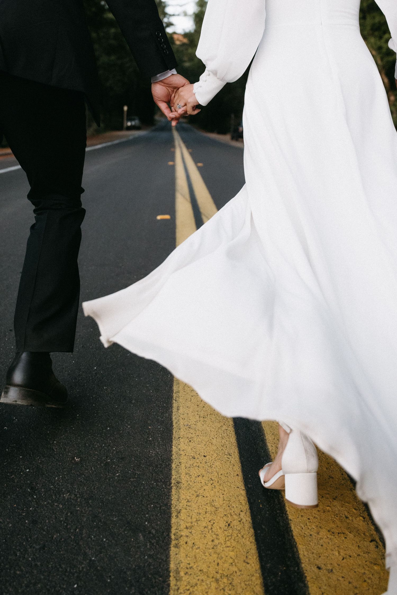 Close-up of bride in white gown and groom in black suit holding hands while walking along a road with yellow center lines.