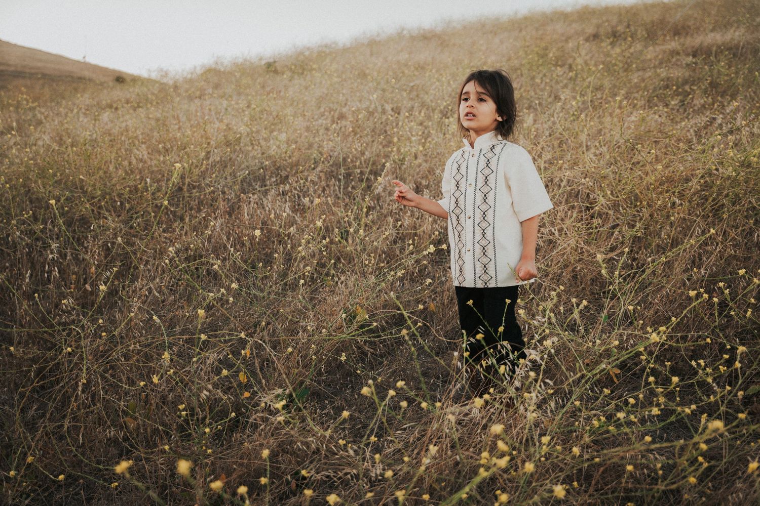 A young child stands in a golden field of wildflowers, wearing a white embroidered shirt and black pants.