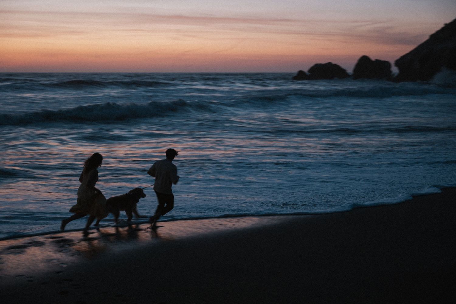 Two people and a dog run along a scenic beach shoreline at sunset.