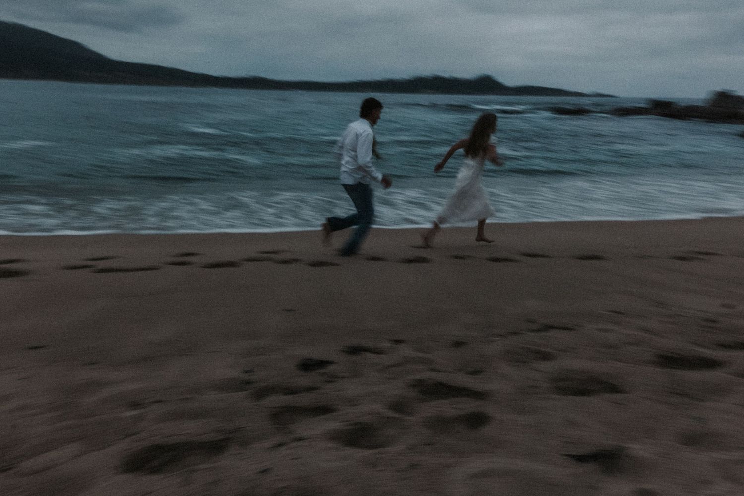 Couple running together on a sandy beach at dusk with ocean waves in the background.Photography by The Y.K. Studio.