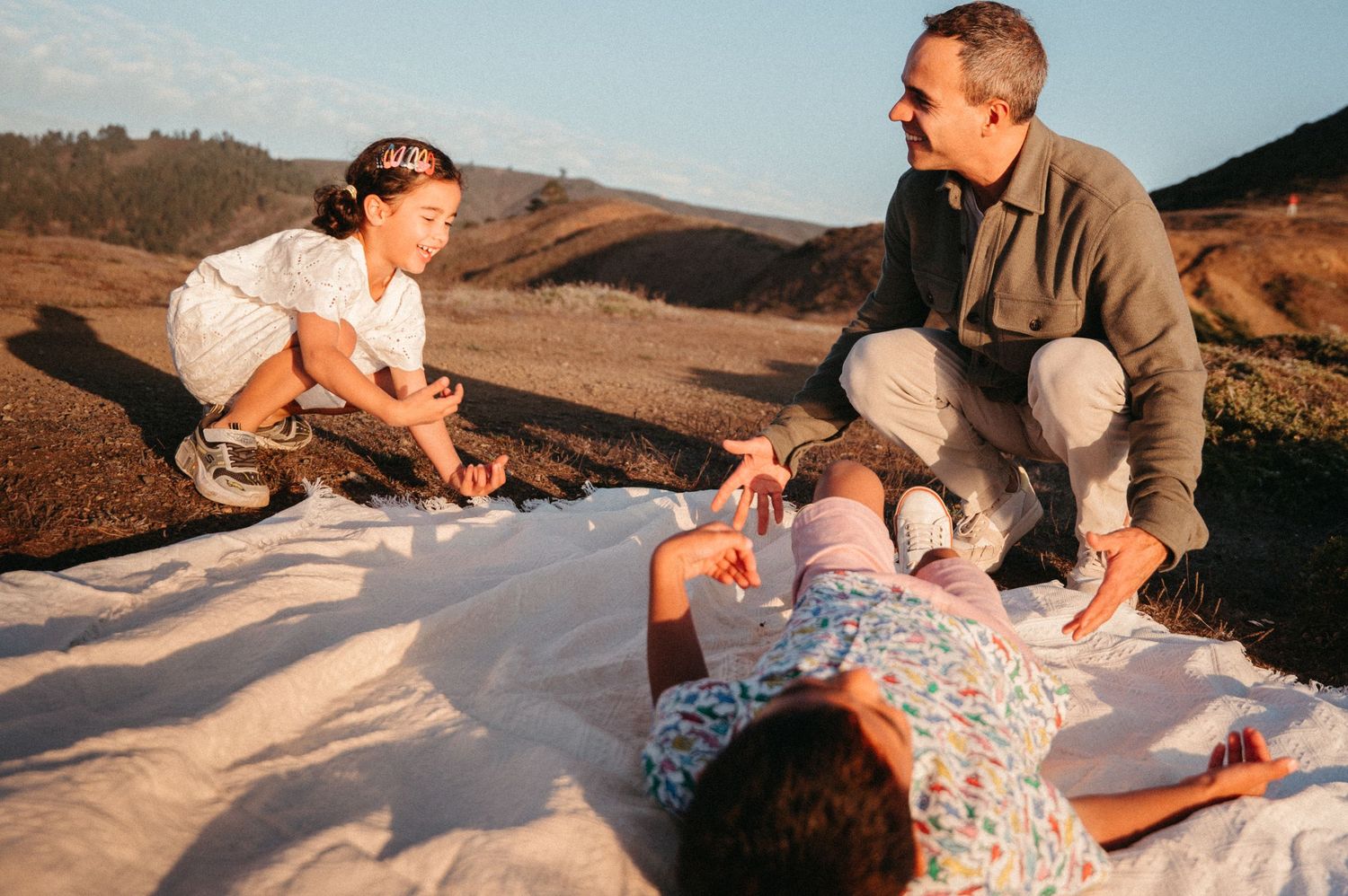 Family enjoying a sunny outdoor picnic on a white blanket in a scenic hillside landscape.
