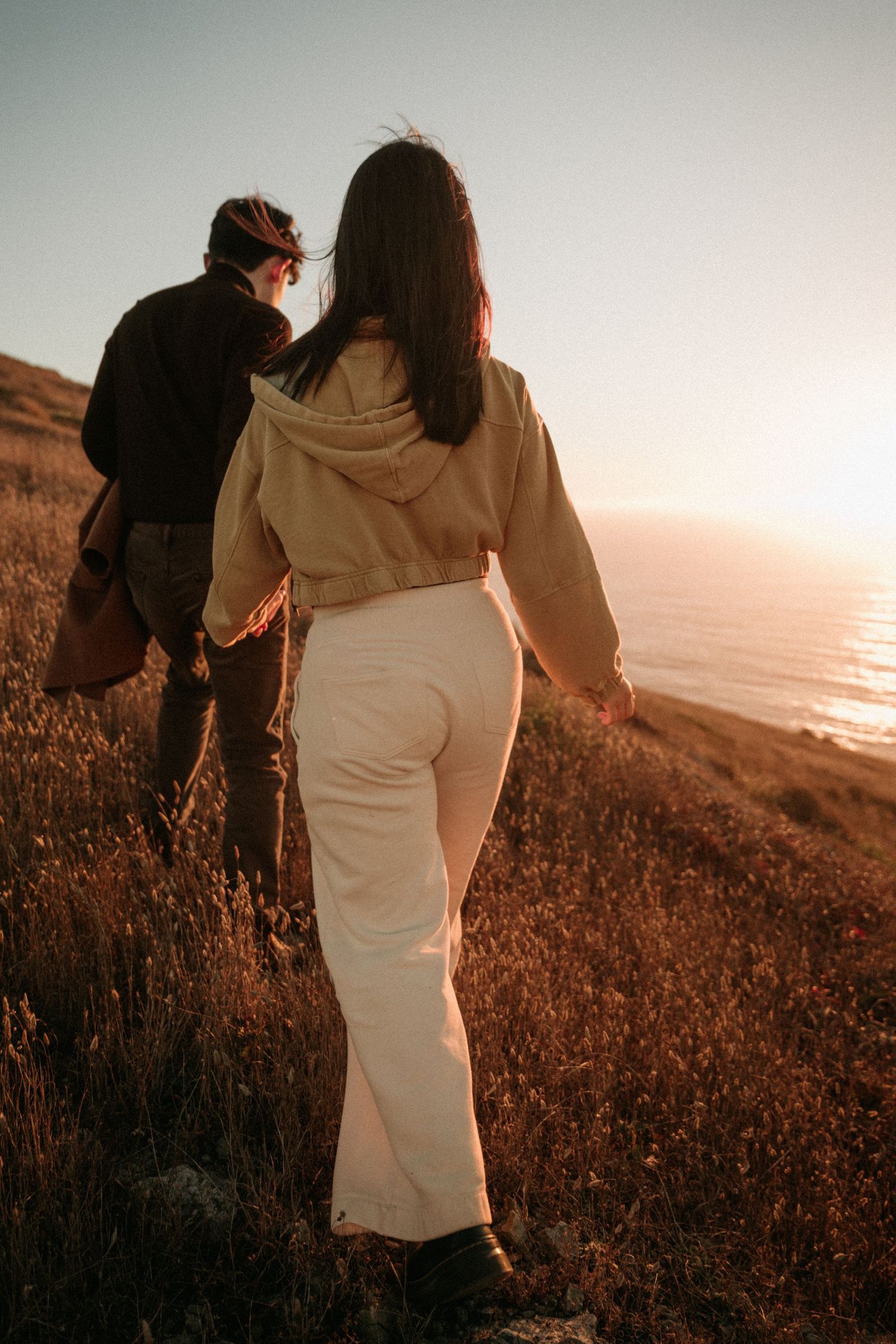 Two people walking hand in hand up a grassy coastal hillside toward the glowing ocean at sunset.