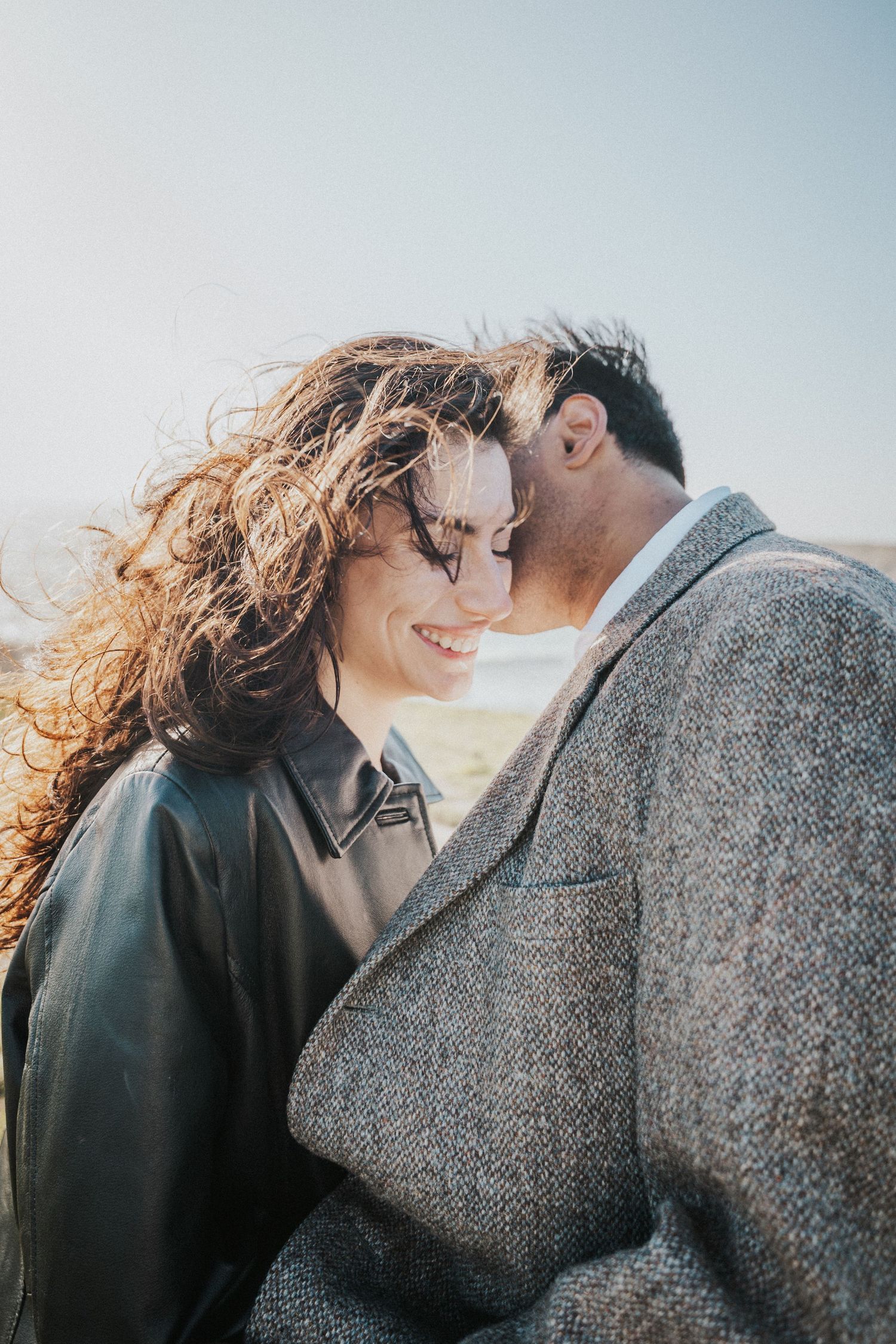 A smiling woman with curly hair and sunglasses shares a romantic moment with a man in a tweed jacket outdoors.