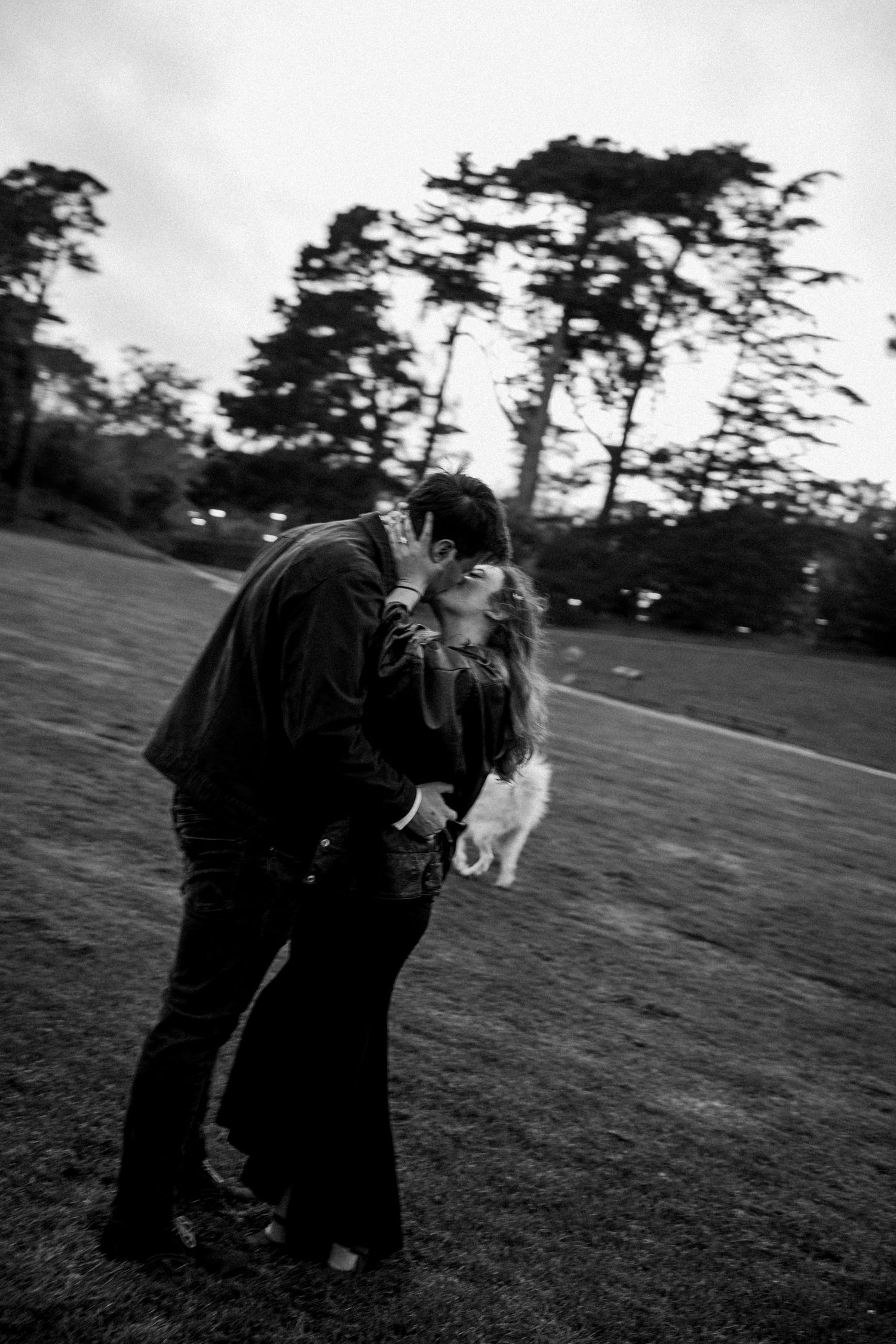 Black and white photo of a couple sharing a romantic kiss outdoors in a park with trees in the background.Photography by The Y.K. Studio.