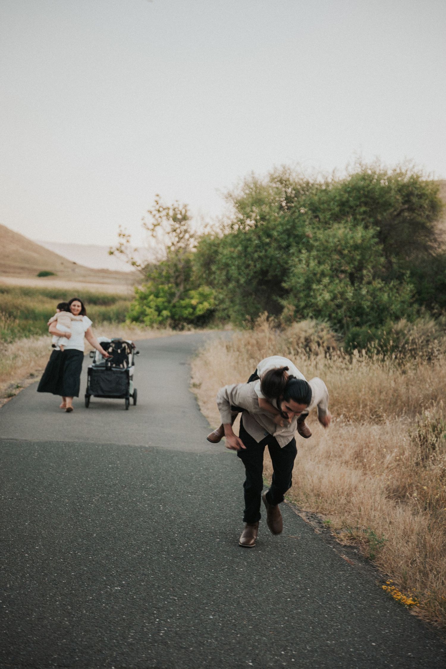 Family enjoys outdoor walk, man giving piggyback ride on path while woman pushes stroller through scenic countryside.