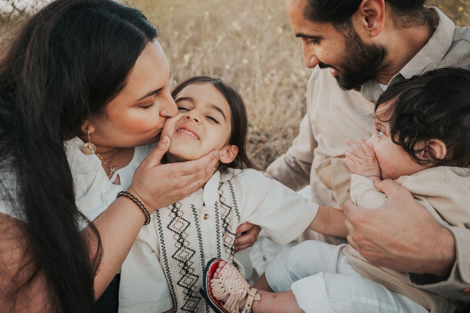 A loving family shares a tender moment outdoors, with a mother kissing her child's cheek while the father holds a baby.