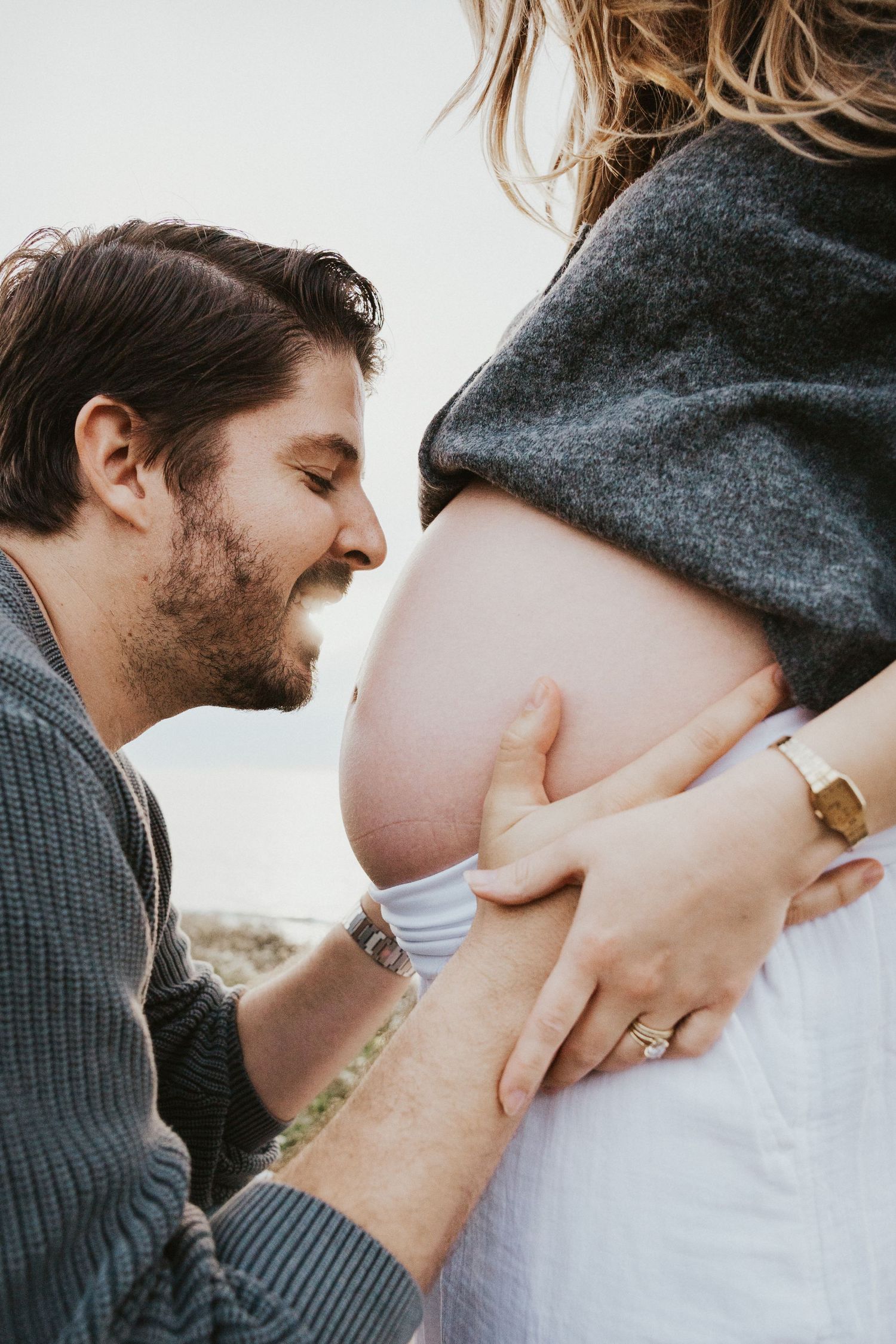 A man smiles while touching a pregnant woman's bare belly outdoors, capturing a tender maternity moment.