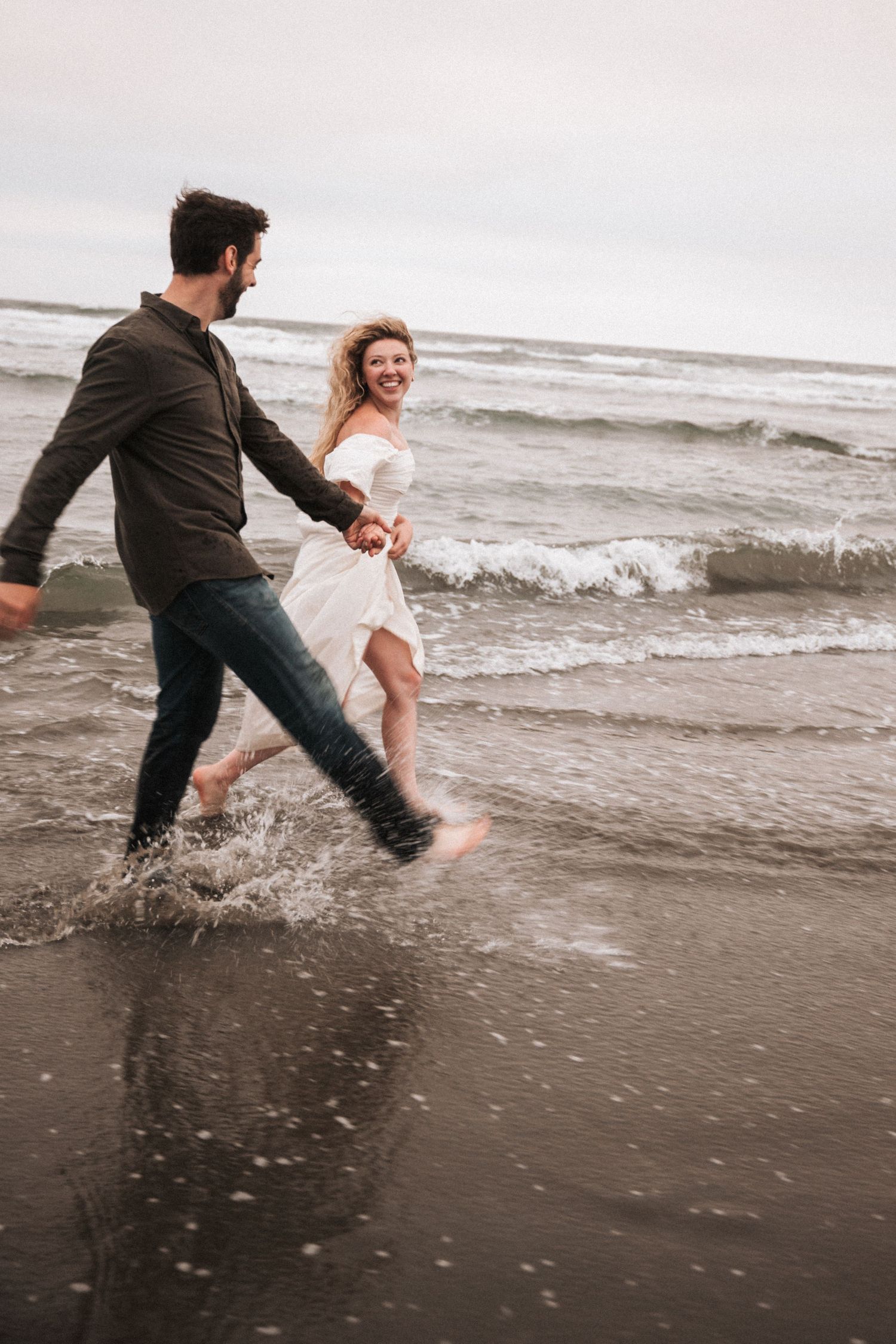 Joyful couple holding hands splashing through ocean waves on a cloudy beach day.