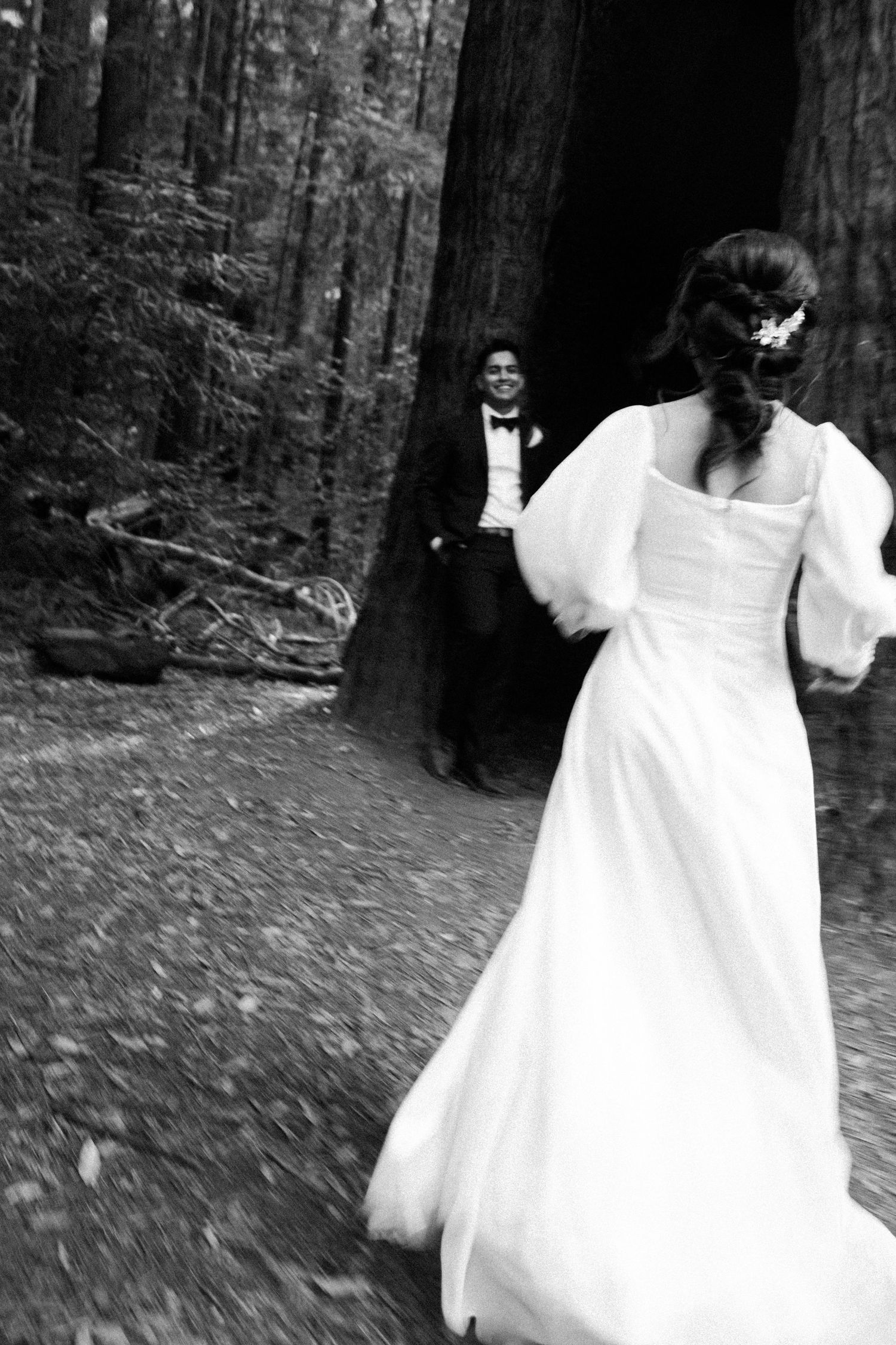 Black and white photo of a bride in a white gown running toward a groom in a tuxedo in a forest wedding.