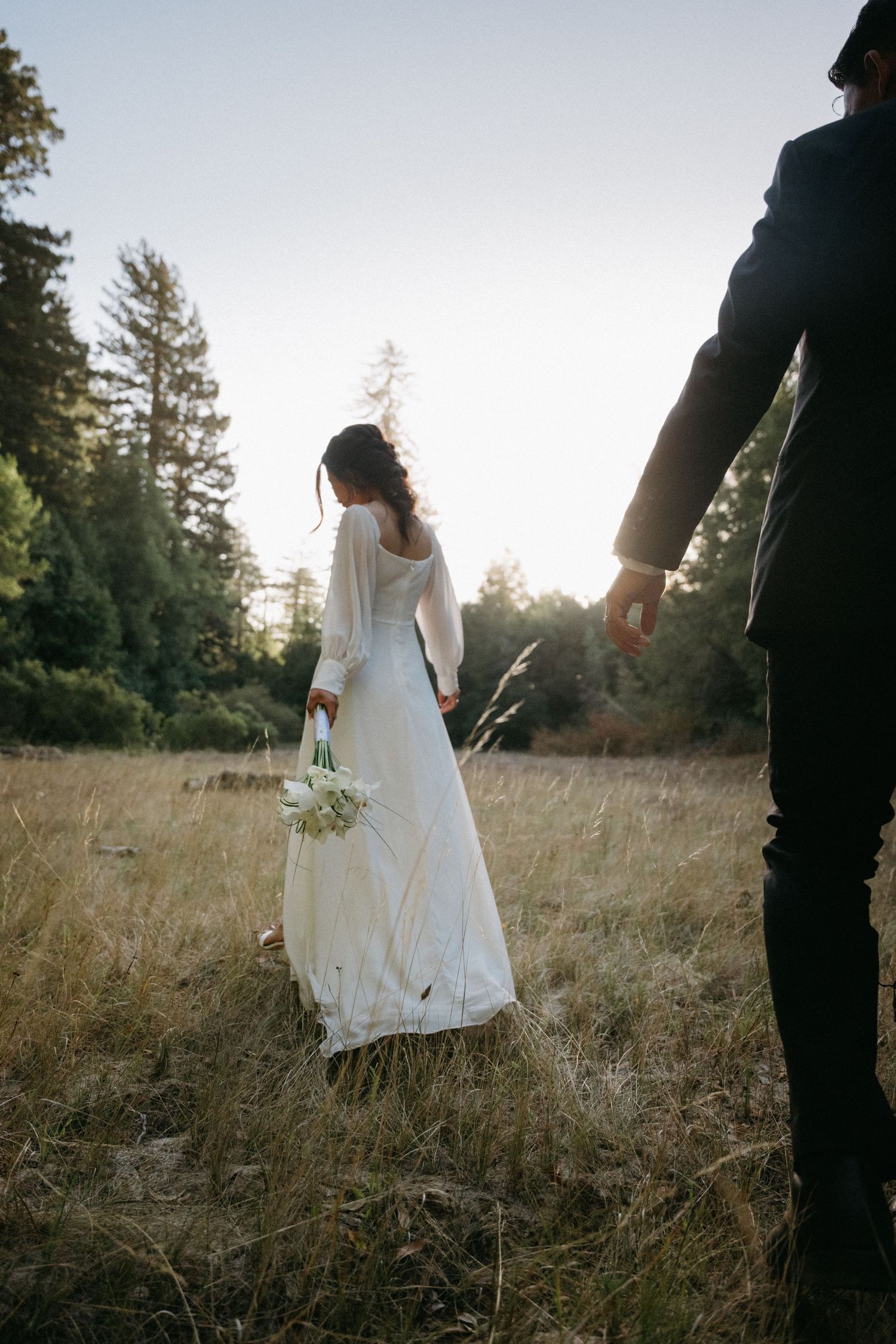 Bride in a long-sleeve white gown holds a bouquet while walking through a golden meadow at sunset with groom following.