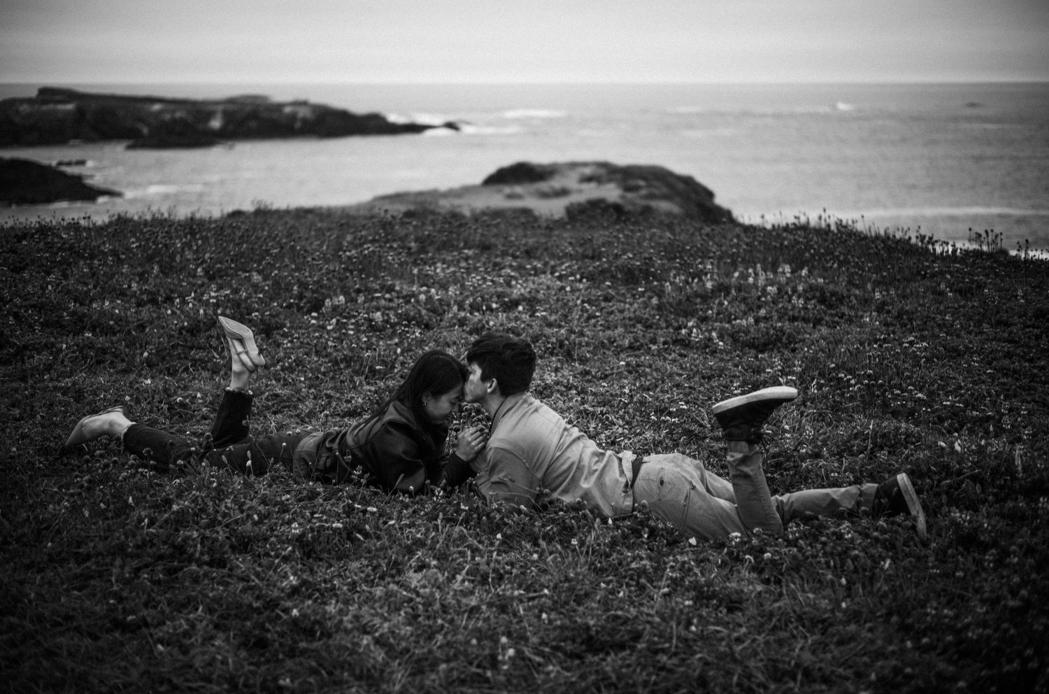 Couple sharing a kiss while lying on a grassy coastal hillside in black and white.