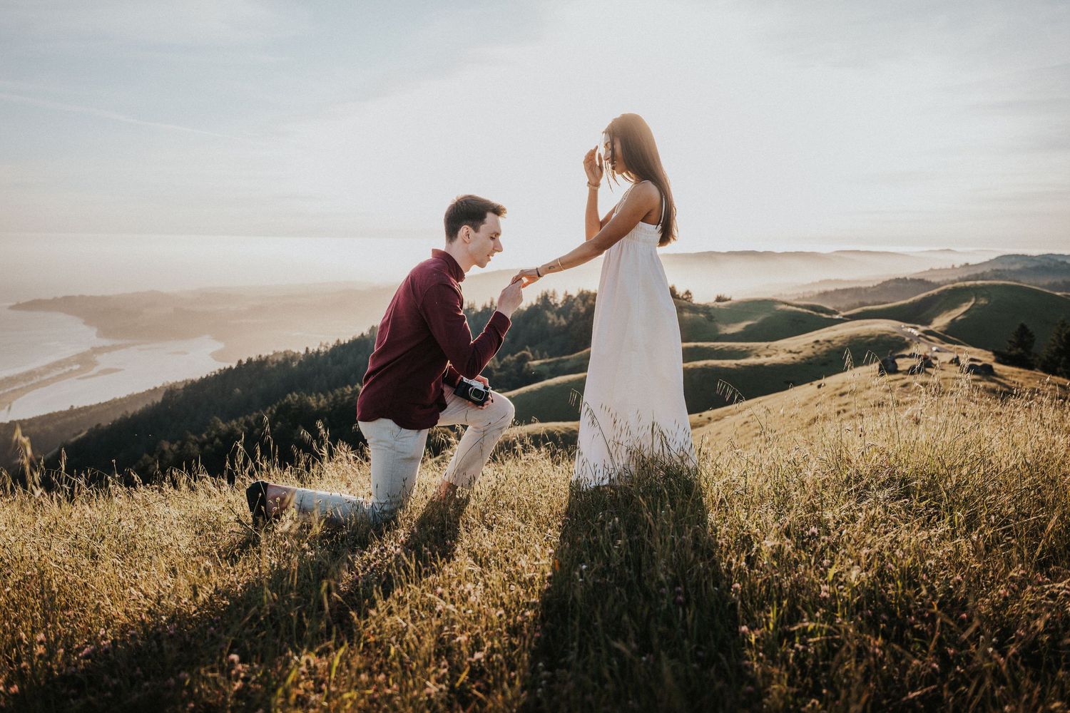 Man proposing to woman on golden hilltop at sunset with scenic coastal landscape in background.