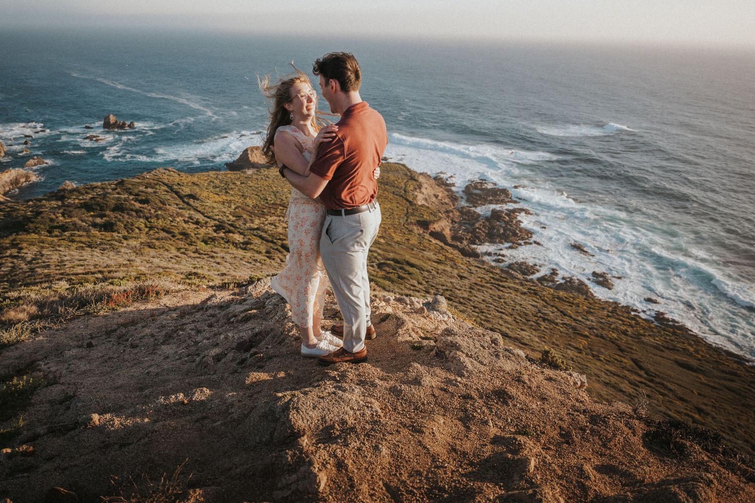 Couple embracing on rocky coastal cliff overlooking ocean waves at golden hour sunset.