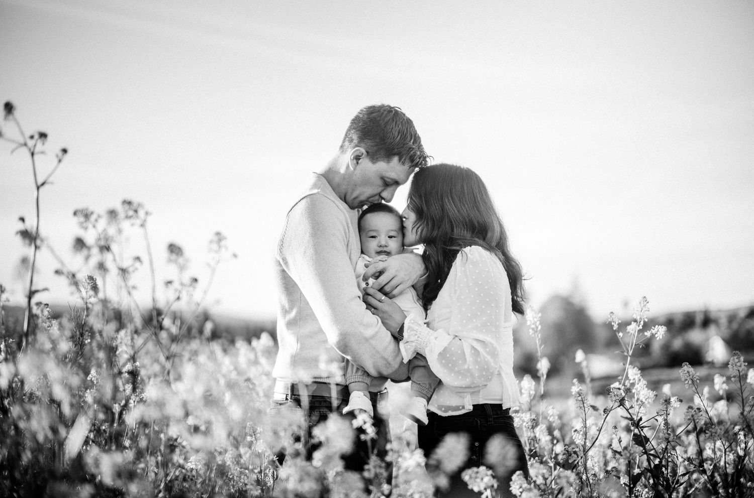 Black and white photo of a couple sharing a tender moment with their baby in a blooming flower field.