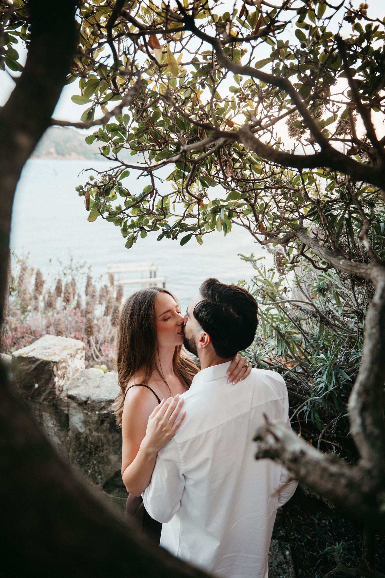 Couple sharing a romantic kiss under a lush tree by the waterside at golden hour.