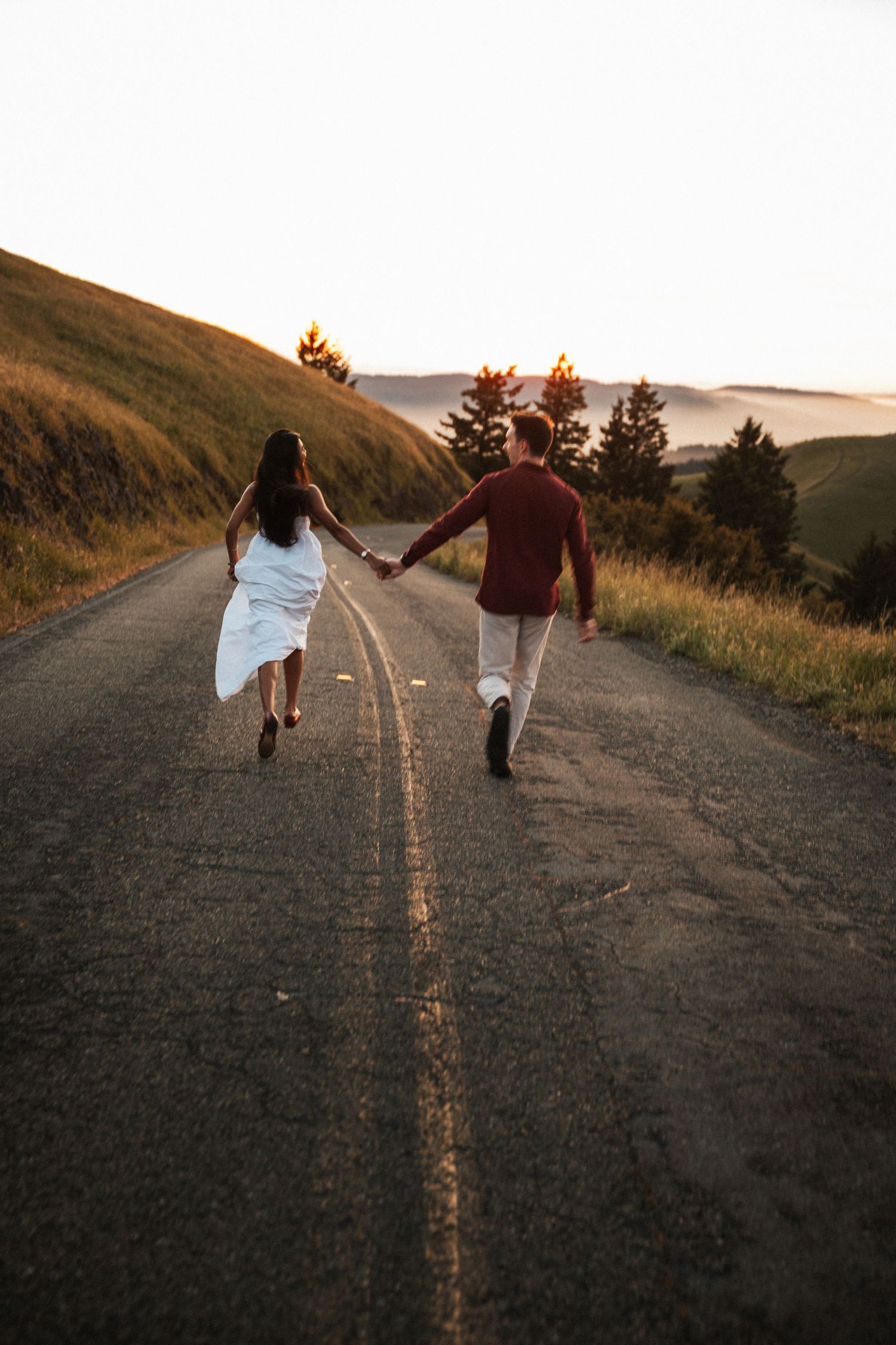 Couple holding hands running down a scenic road at sunset, surrounded by rolling green hills and pine trees.