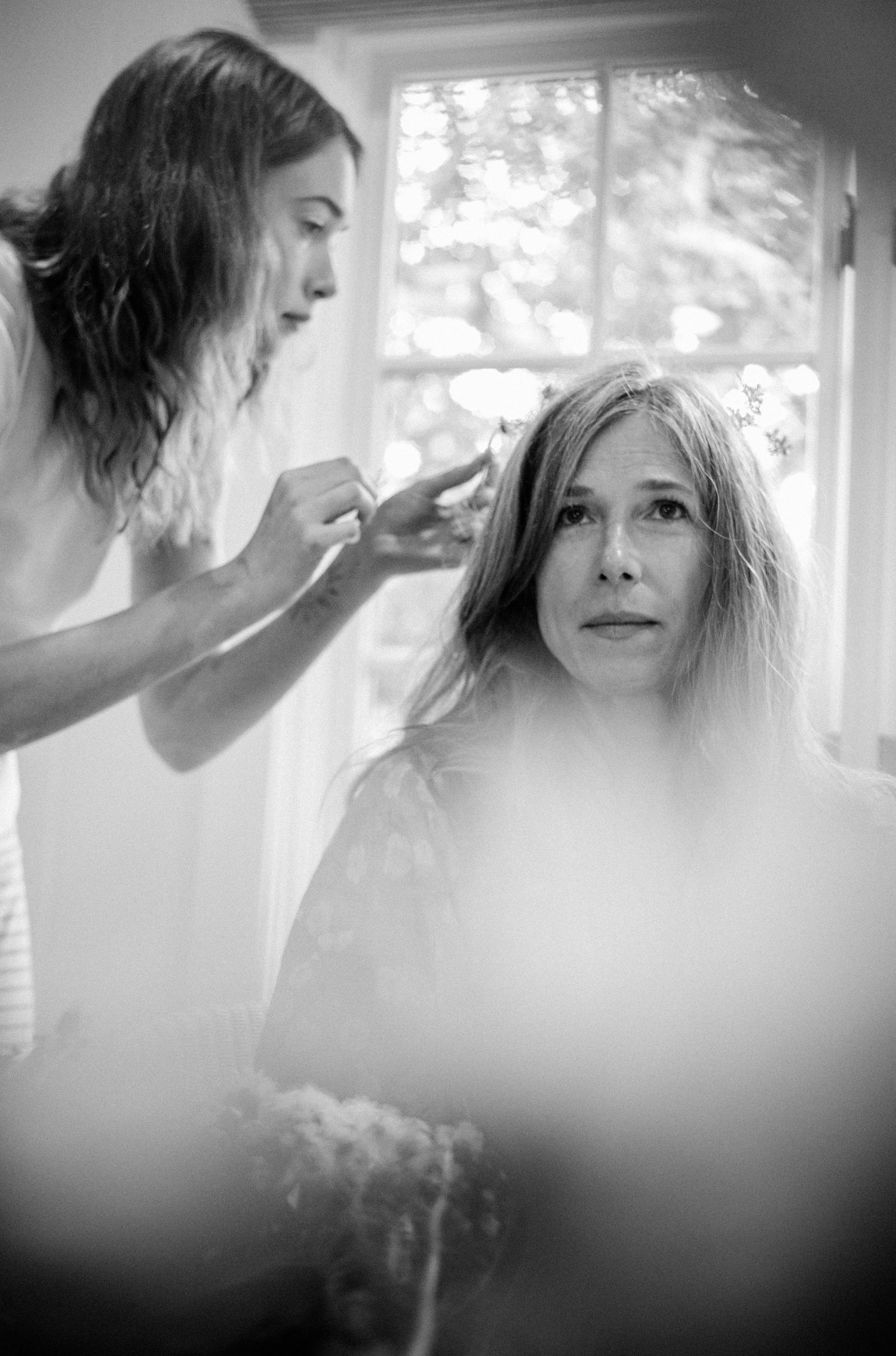 Black and white photo of a woman getting her hair styled by a hairdresser near a bright window.