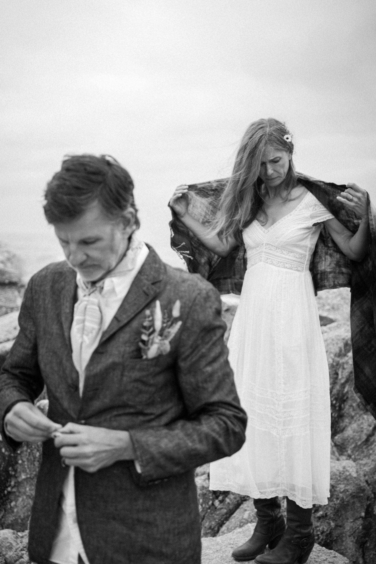 A bride and groom in black and white photo on rocky shore, she holds his jacket.