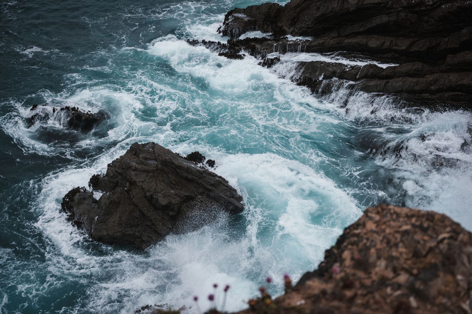 Dramatic ocean waves crashing against rocky coastal cliffs with turquoise blue water swirling below.