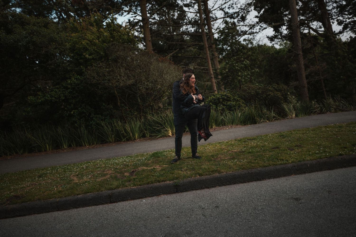 Couple embracing on a grassy roadside path surrounded by tall green trees at dusk.