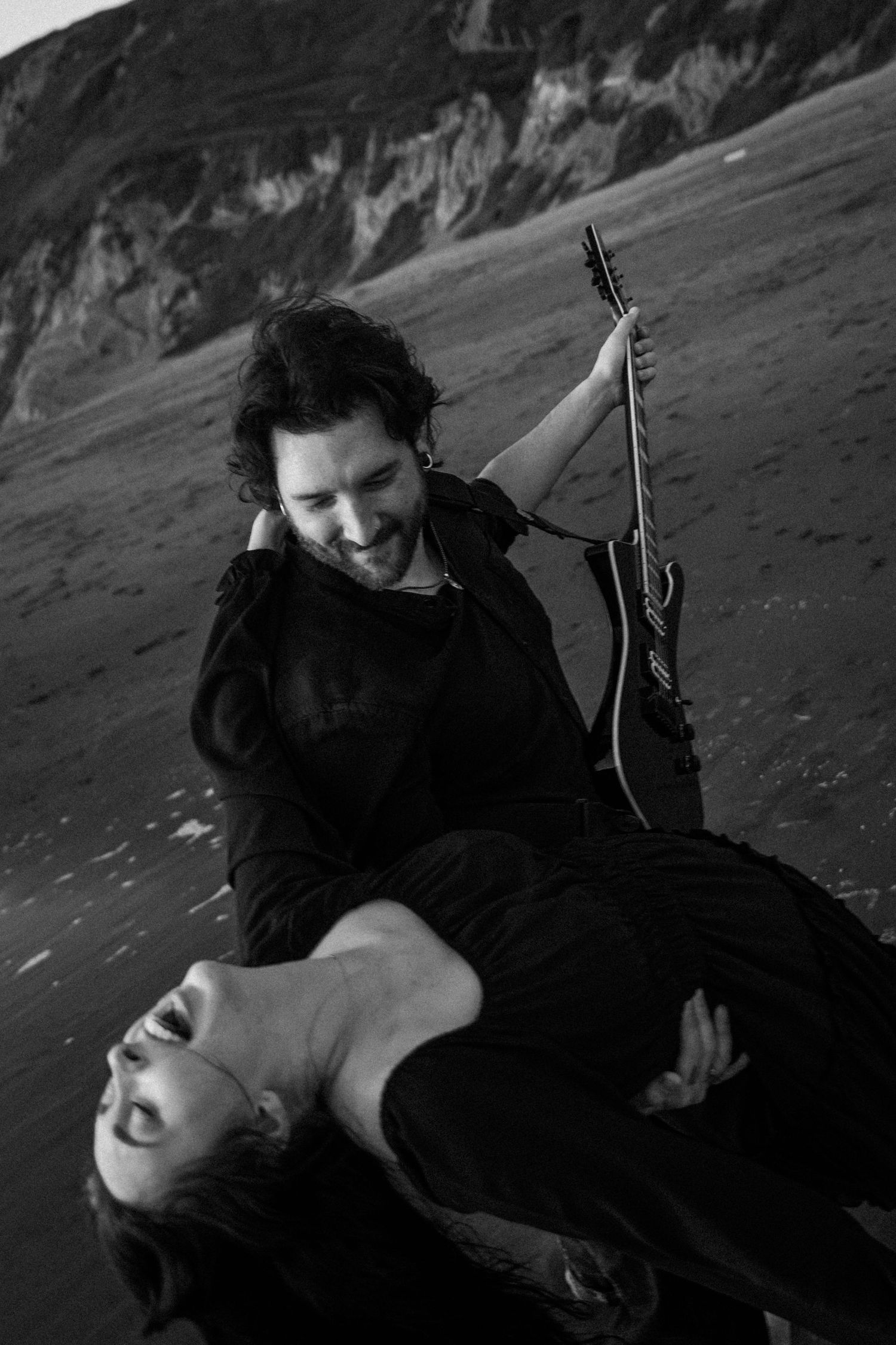 Black and white photo of a laughing couple on a beach, man holding guitar while dipping woman dramatically.