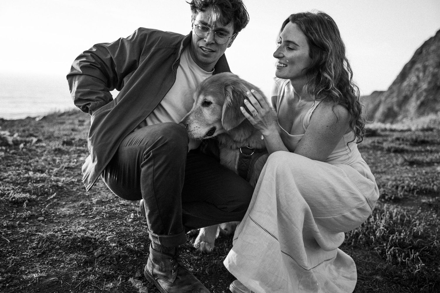 Black and white photo of a couple in wedding attire petting a golden retriever dog outdoors near coastal cliffs.