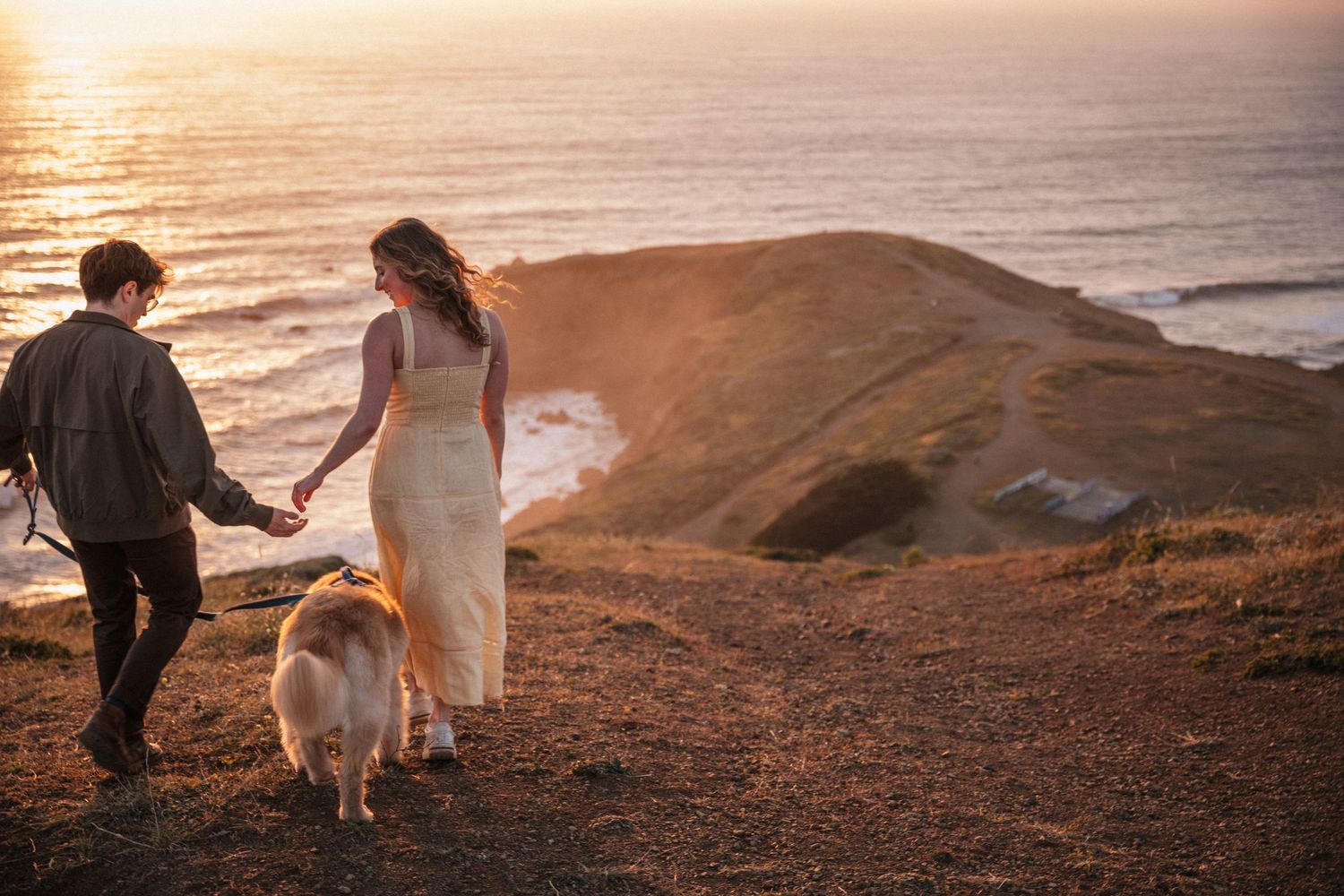 Couple walks hand in hand with golden dog along coastal hillside trail at golden hour sunset.